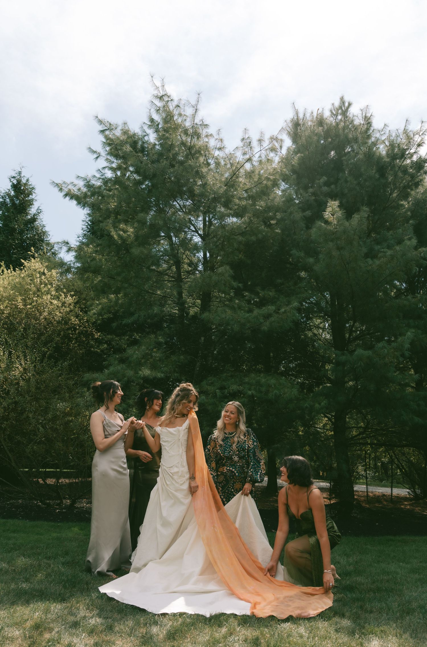 Group of women in formal dresses walking together on a grassy lawn with trees in the background.