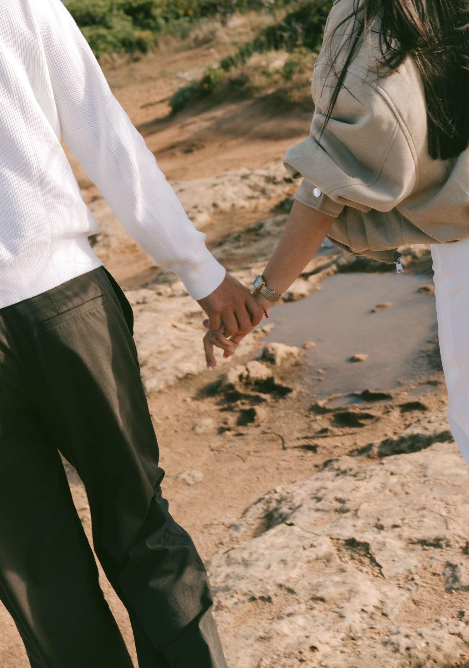 Couple holding hands on a rocky beach, wearing casual white and gray outfits, with sandy terrain in the background.