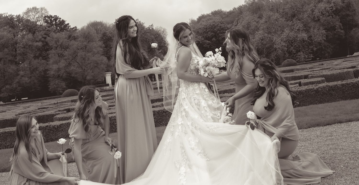 Wedding party in sage green dresses assists with bride's dress on grassy hillside in Germany.