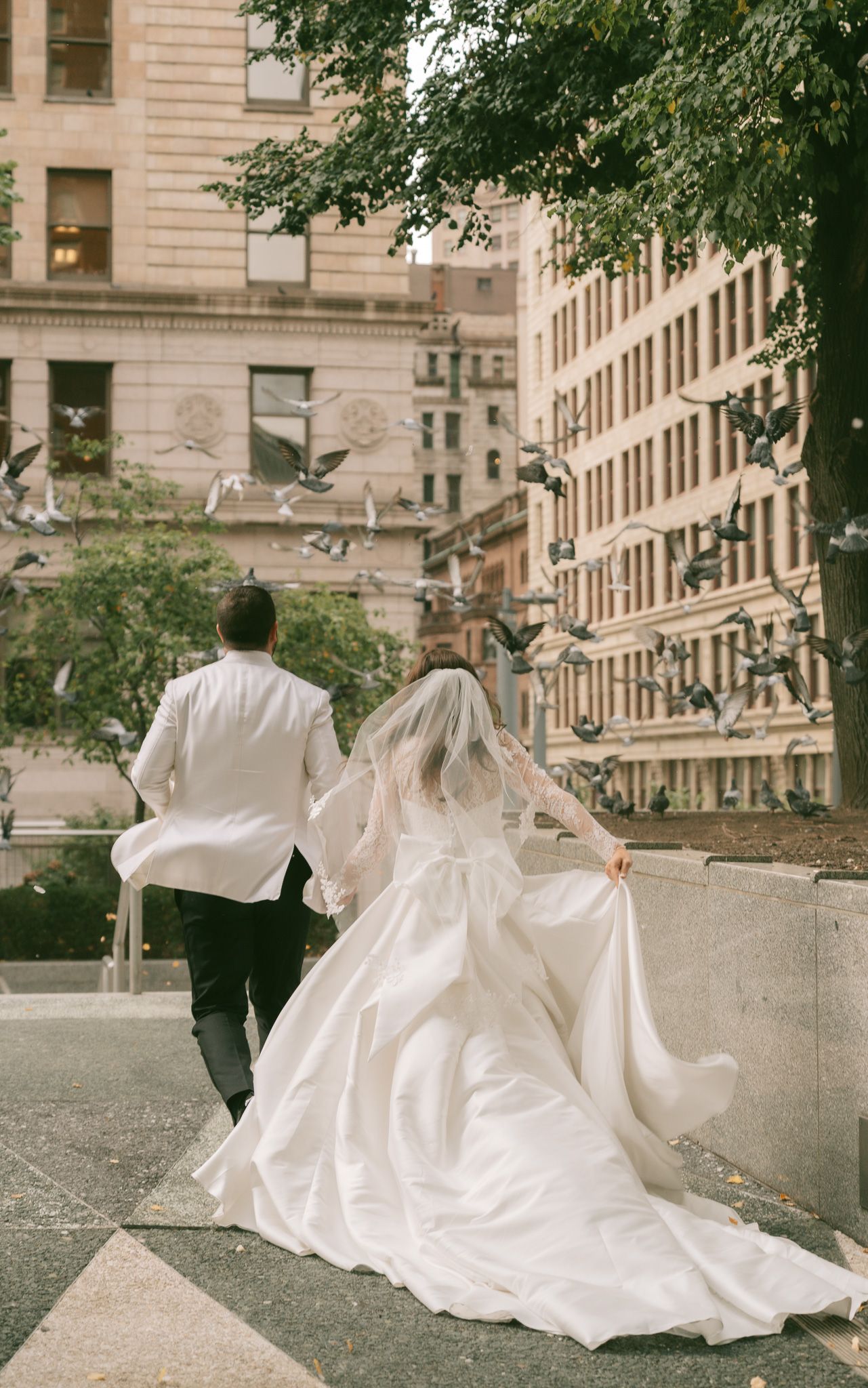A flowing white wedding dress trails behind a couple walking in an urban setting with historic buildings in downtown pittsburgh, pa.
