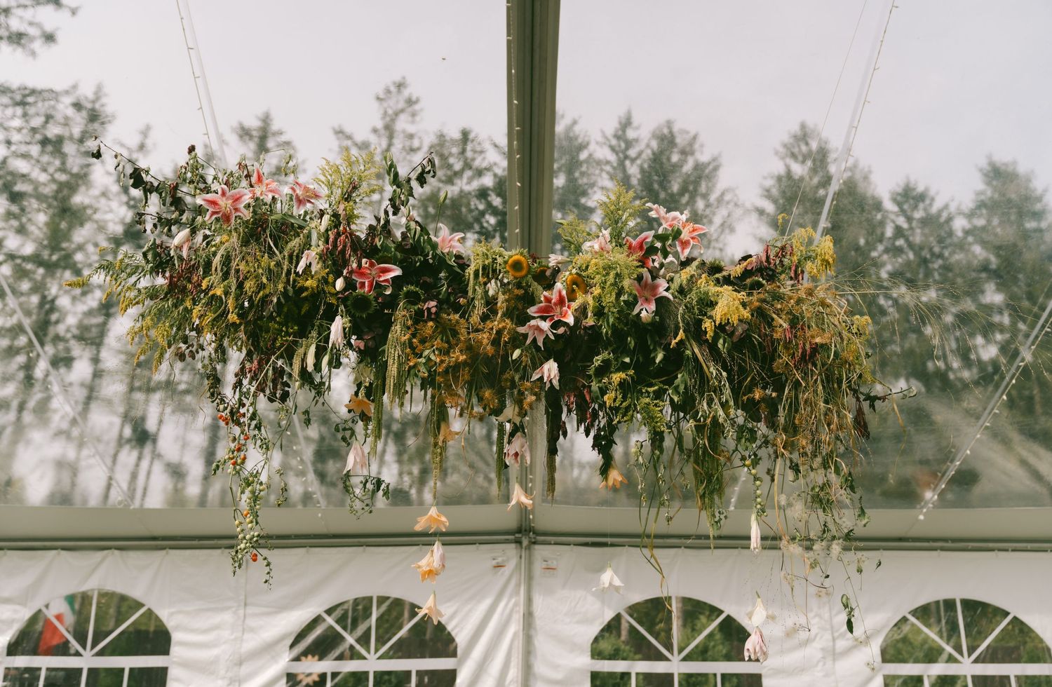 A wedding ceremony setup outdoors with floral arrangements hanging from a wooden pergola against a misty forest backdrop.