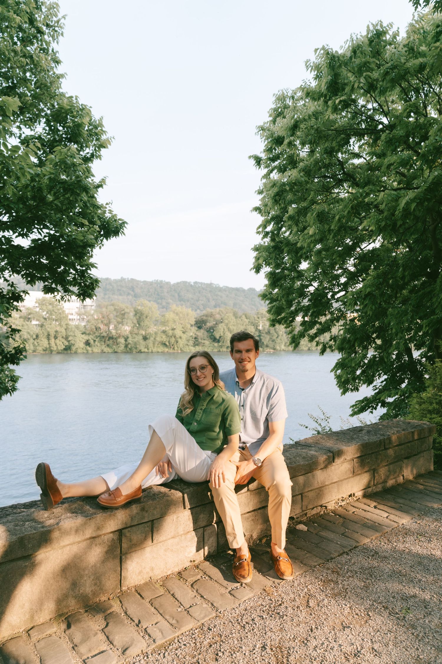 A couple sits together on stone steps overlooking a serene lake surrounded by trees.