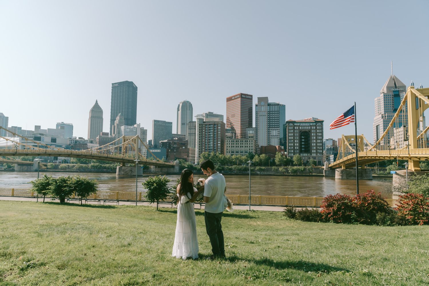 A couple stands on a grassy lawn overlooking the Pittsburgh skyline and yellow bridge on a sunny afternoon in north shore.