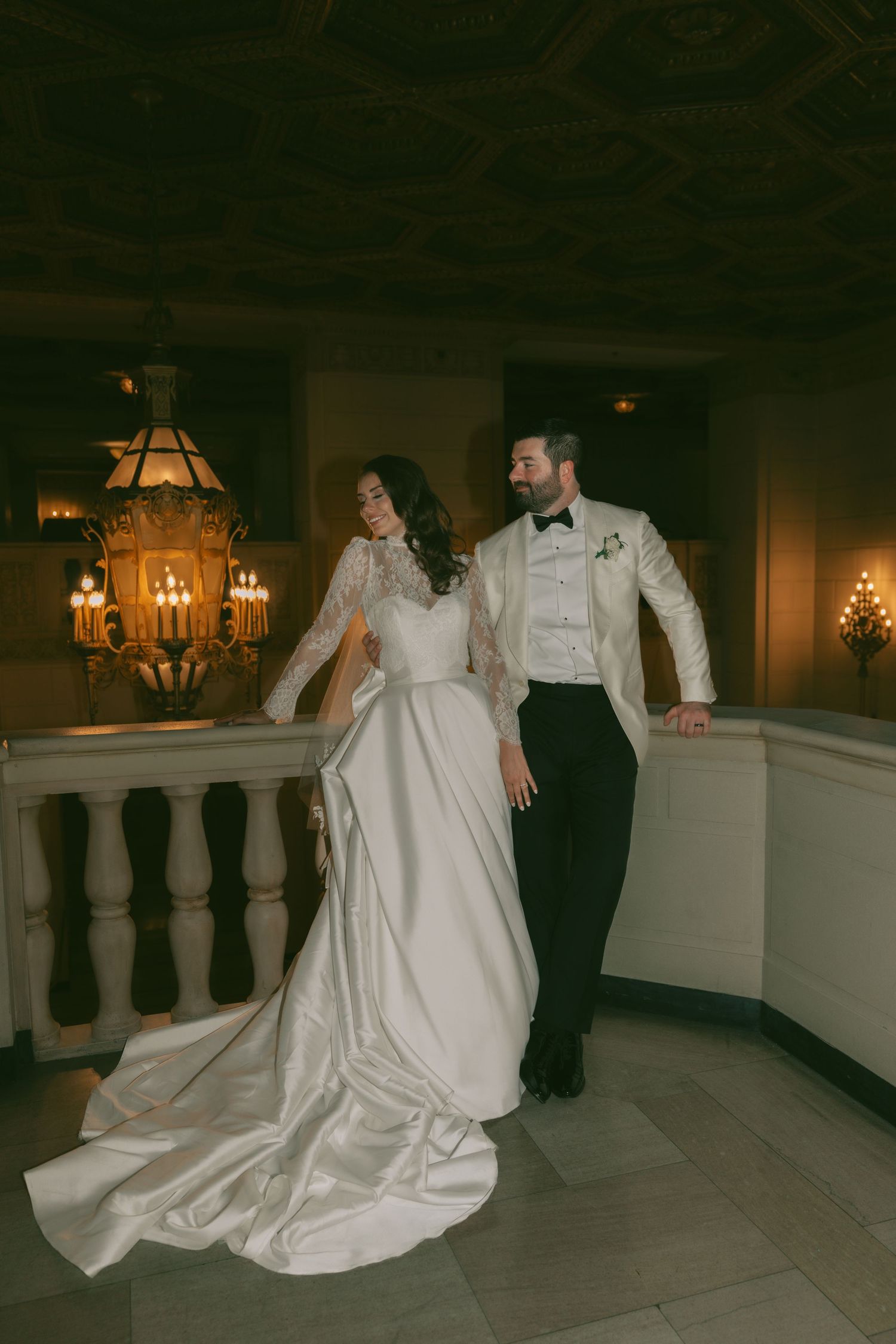A couple in formal wedding attire dances and twirls together in an elegant ballroom with warm lighting and stone balustrades.