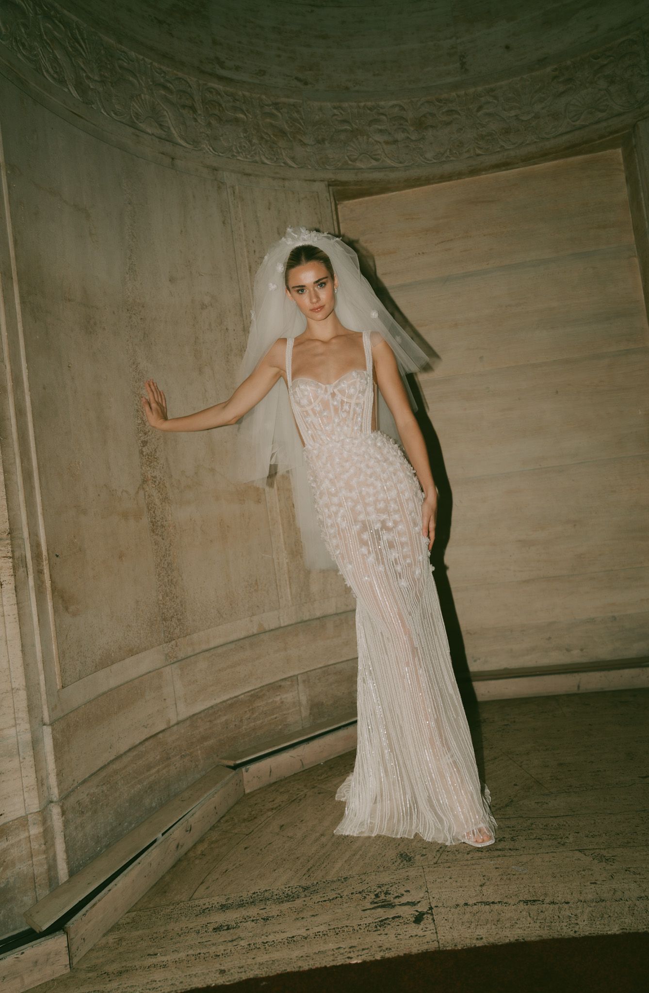 A bride poses in a fitted white lace wedding gown with a flowing veil against a concrete wall.