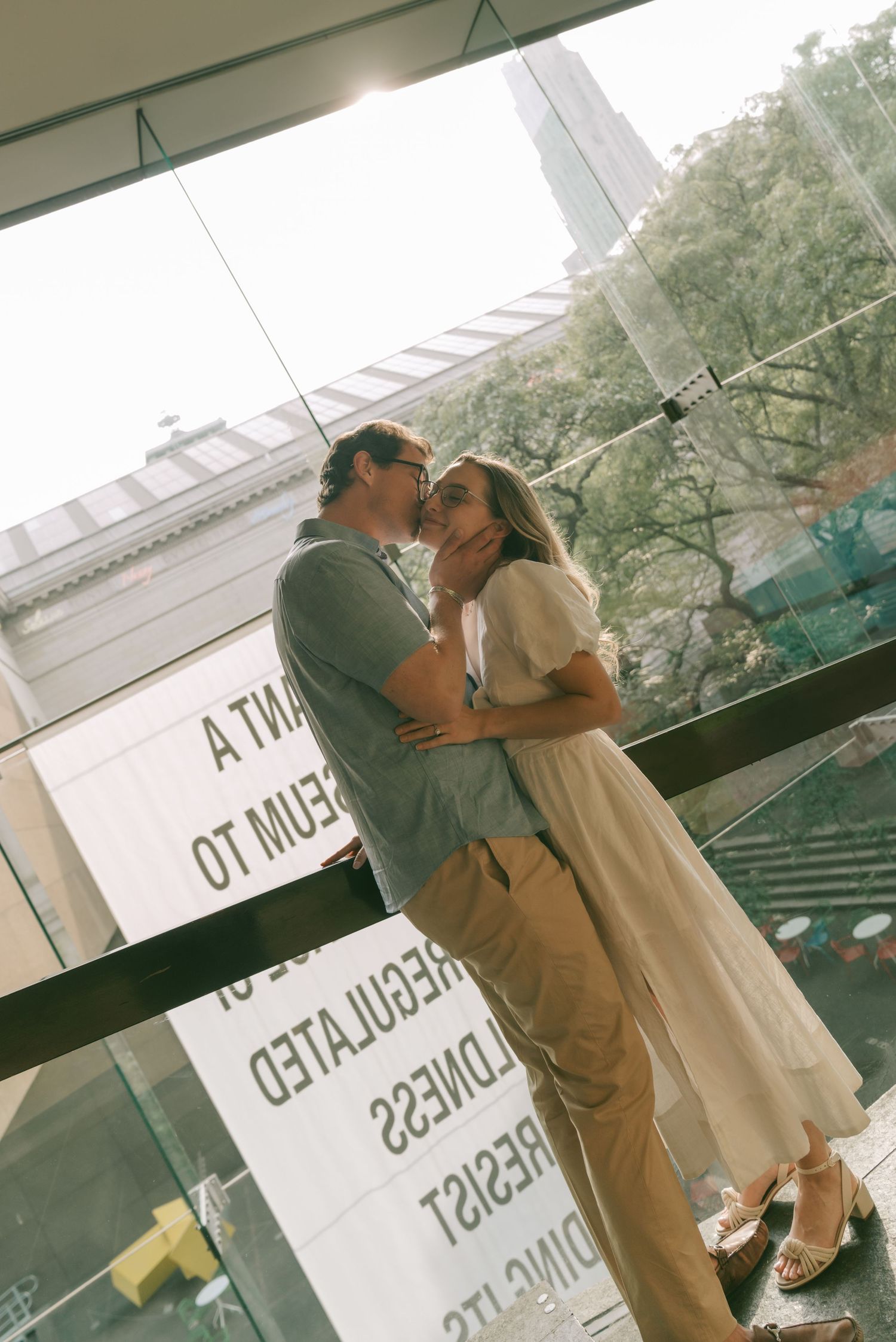 A romantic couple sharing a tender kiss on a balcony in warm sunlight with window reflections visible.