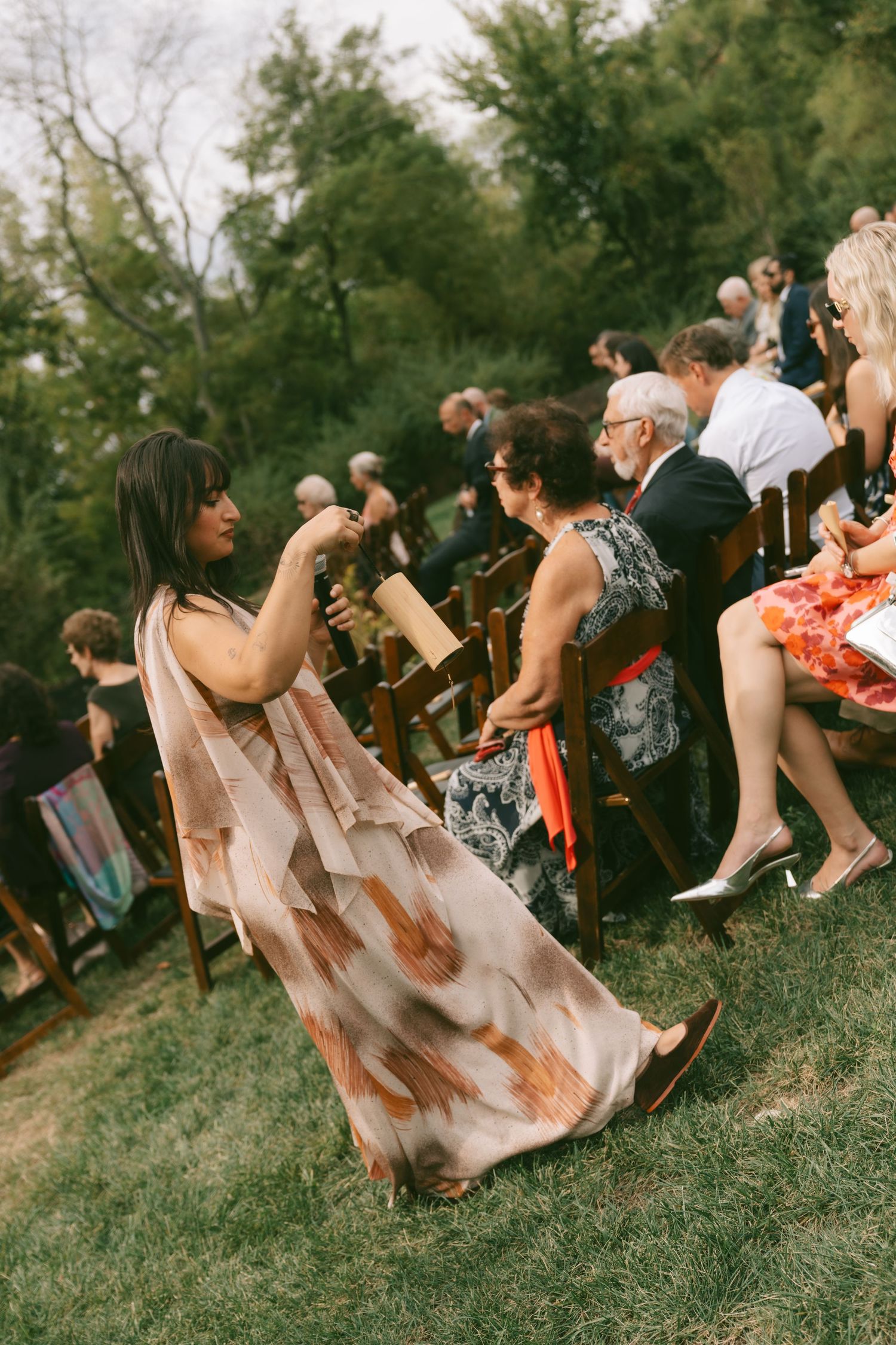Wedding guests seated on wooden chairs at an outdoor ceremony surrounded by greenery.