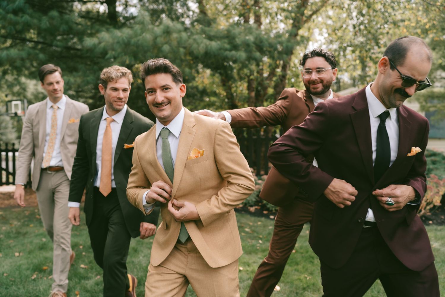 Groomsmen in tan and dark suits laughing together outdoors.