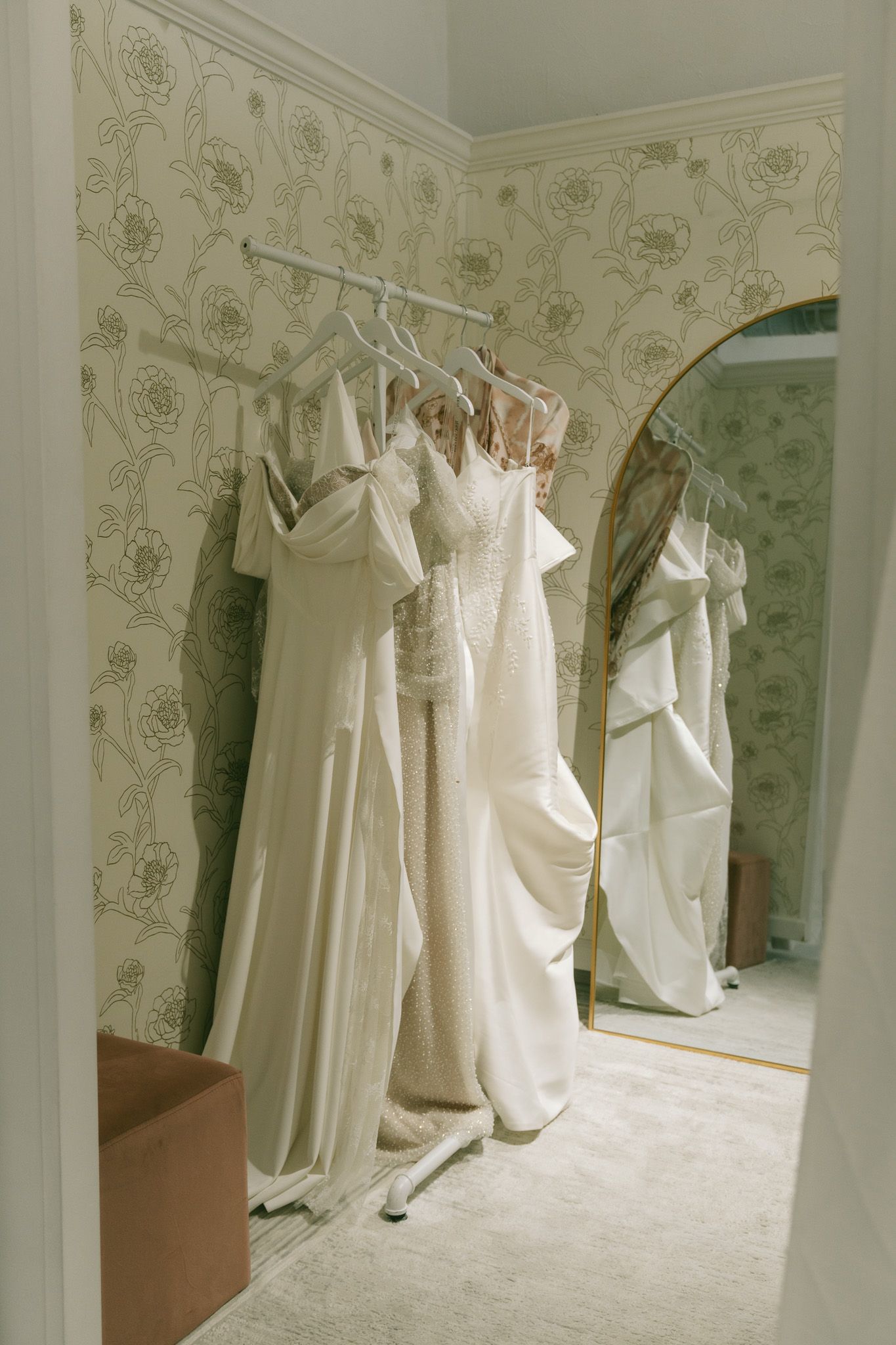 Multiple white wedding dresses displayed on hangers against a floral wallpapered wall in a bridal boutique.