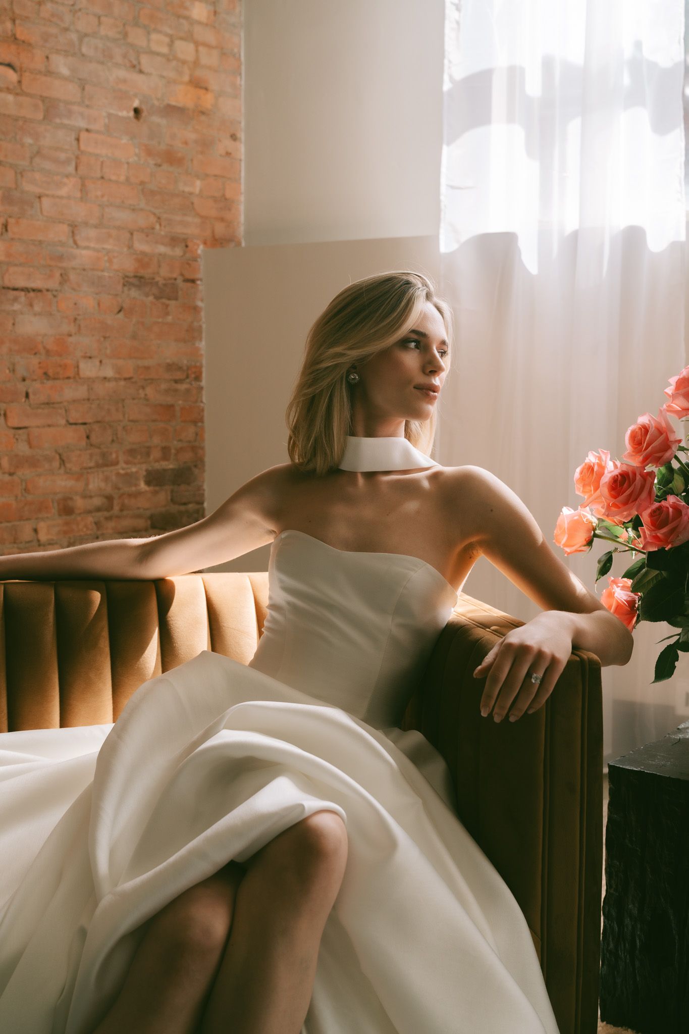 Sun-lit bridal portrait with coral roses against exposed brick in an industrial space.