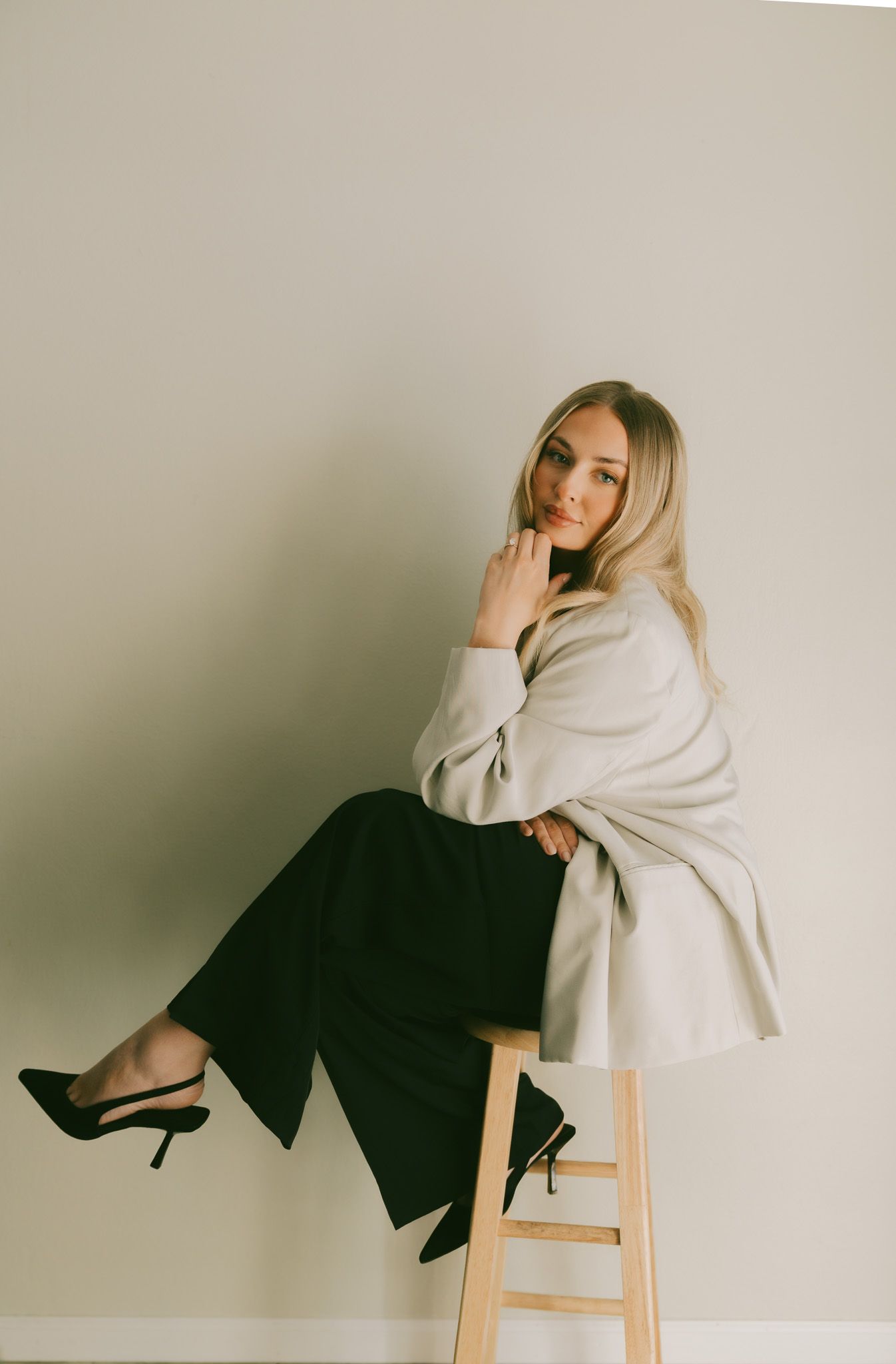 A person in a white sweater and black pants sits pensively on a wooden stool in a minimalist studio setting.