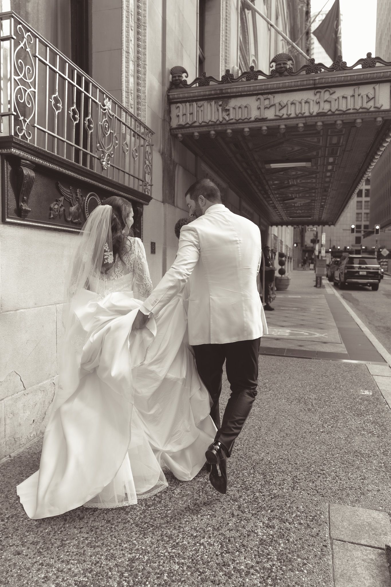 Black and white photo of newlyweds walking along a city sidewalk under an ornate building entrance in downtown pittsburgh, pa.