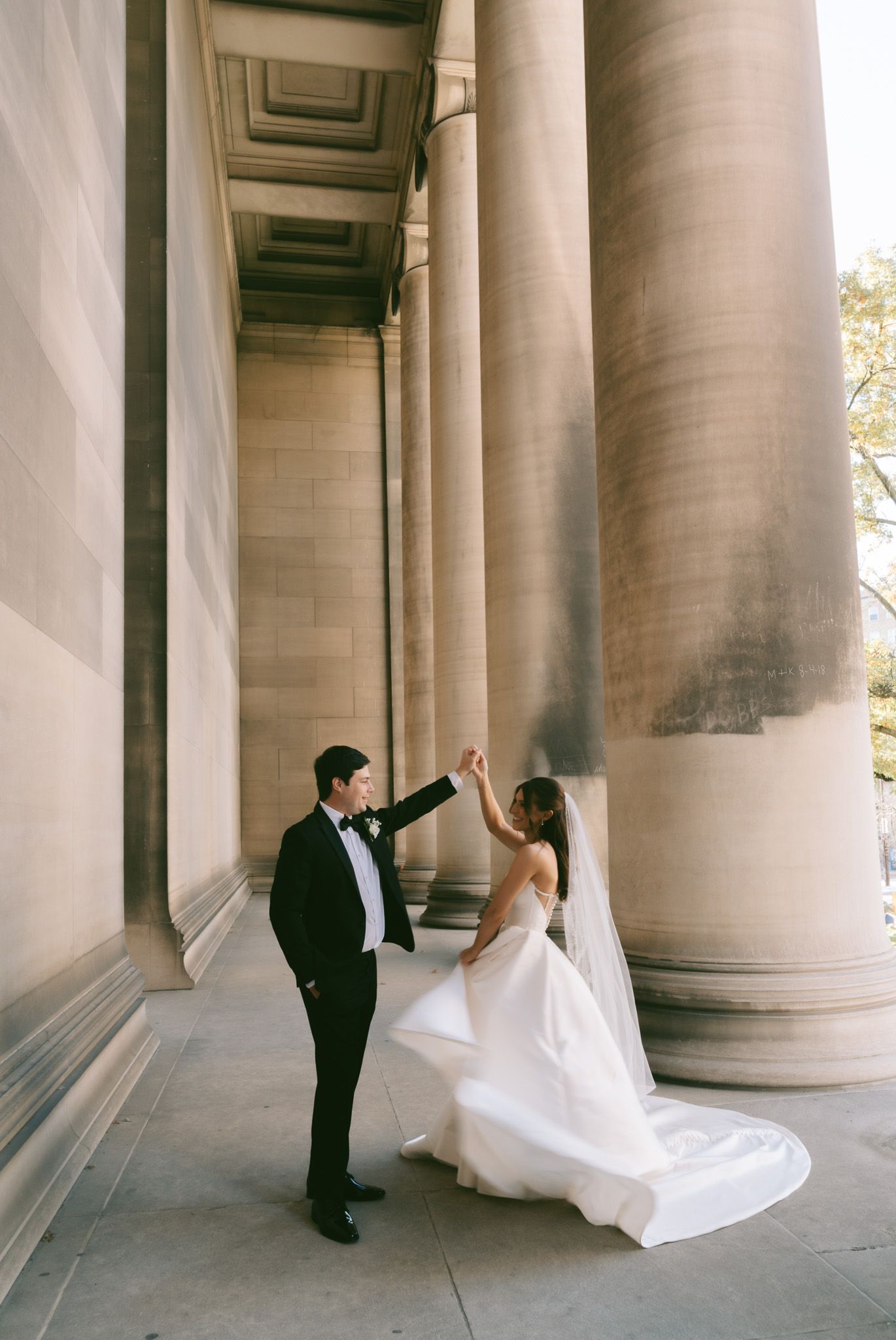 A couple in wedding attire dancing and twirling between grand classical columns of a historic building.