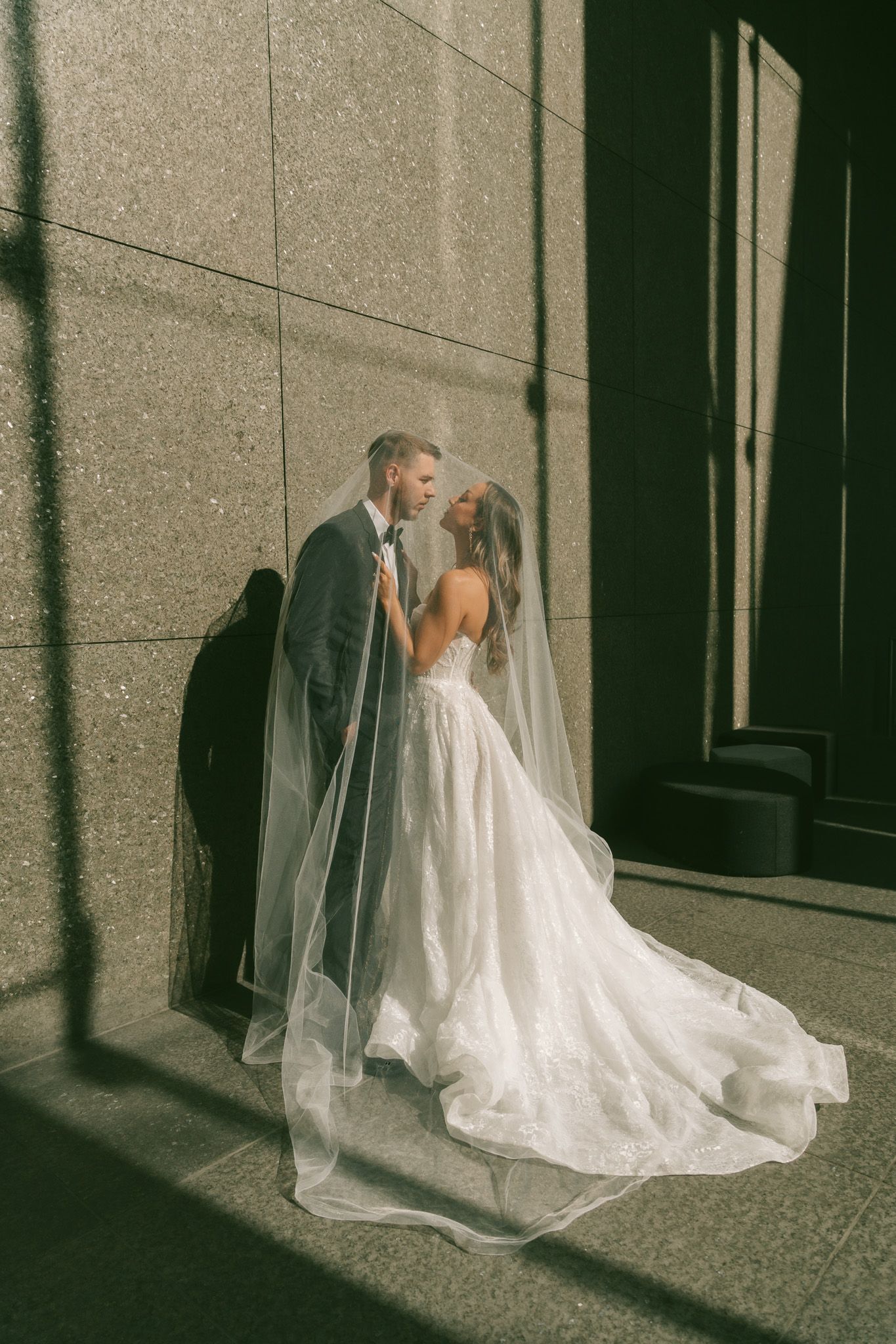 A bride and groom share an intimate kiss against a stone wall as sunlight creates dramatic shadows.