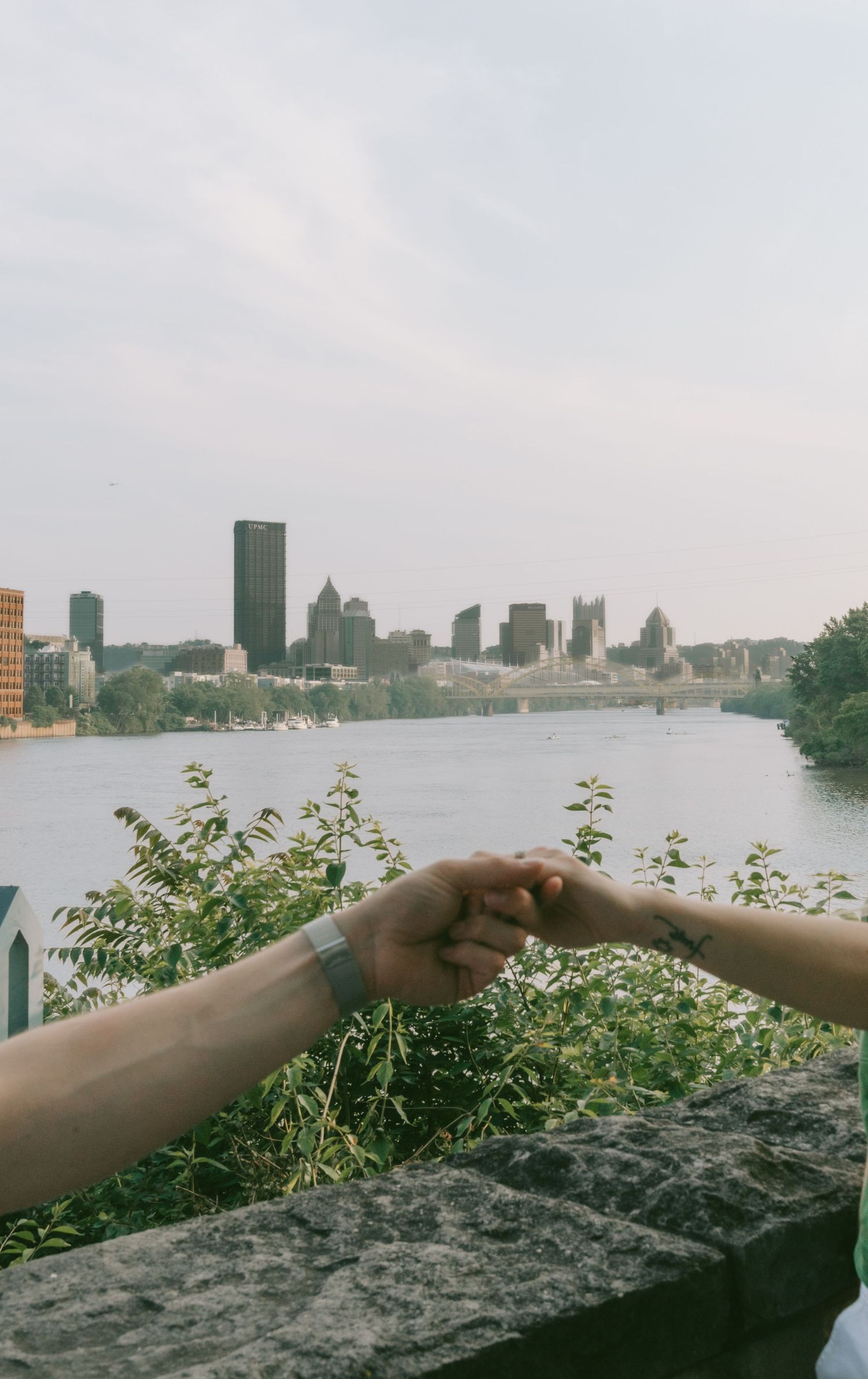 Two hands reach out across a stone wall with a cityscape and river view in the background.