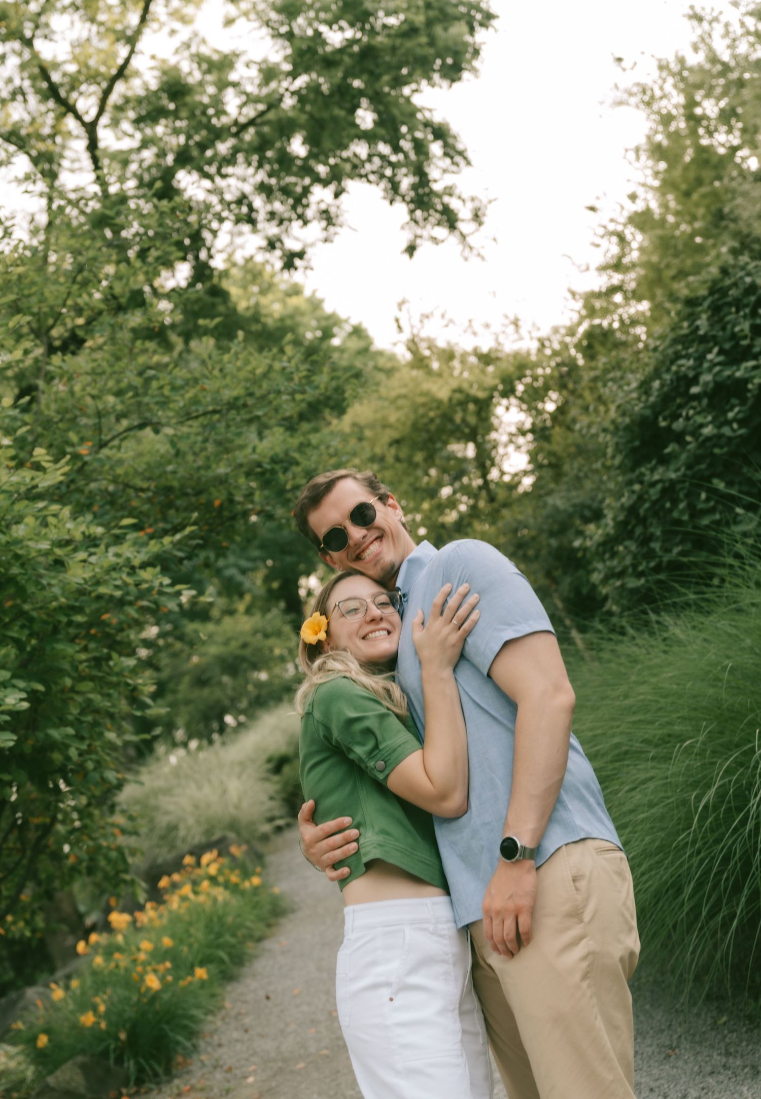 A couple in sunglasses poses playfully on a garden path lined with ornamental grasses and blooming flowers.