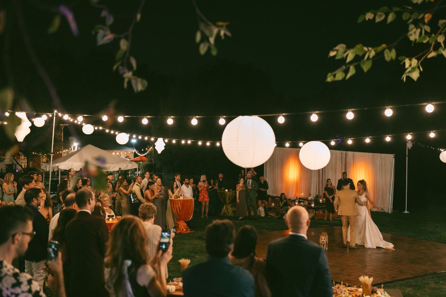 Crowd gathered at outdoor evening celebration under twinkling string lights and white paper lanterns.