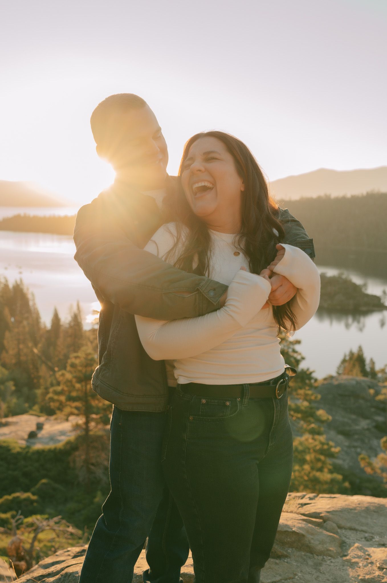 Couple embracing and laughing on a cliff overlooking a lake at sunset.
