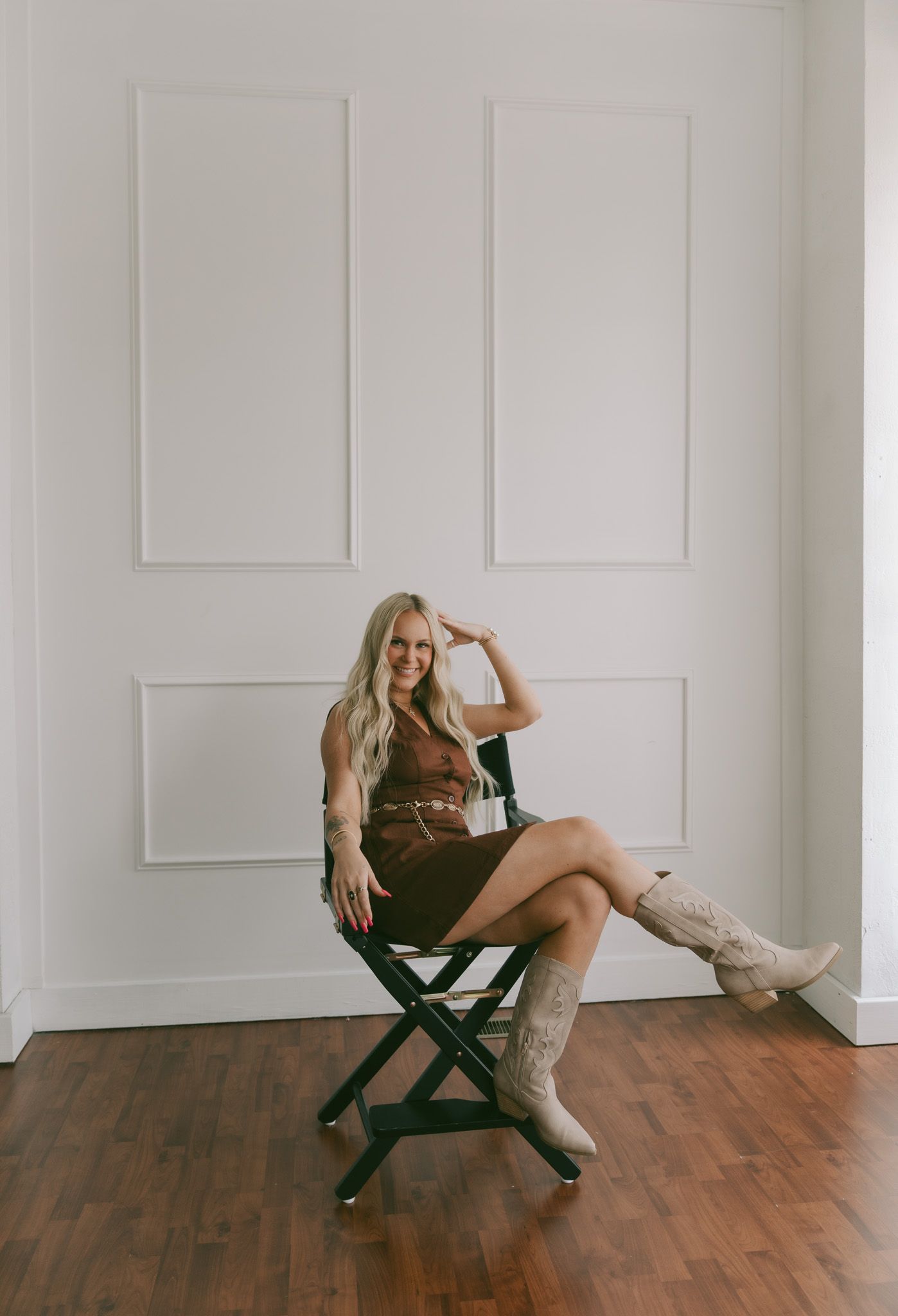 A model poses in a black dress and tall boots while sitting on a green folding chair against a white paneled wall.