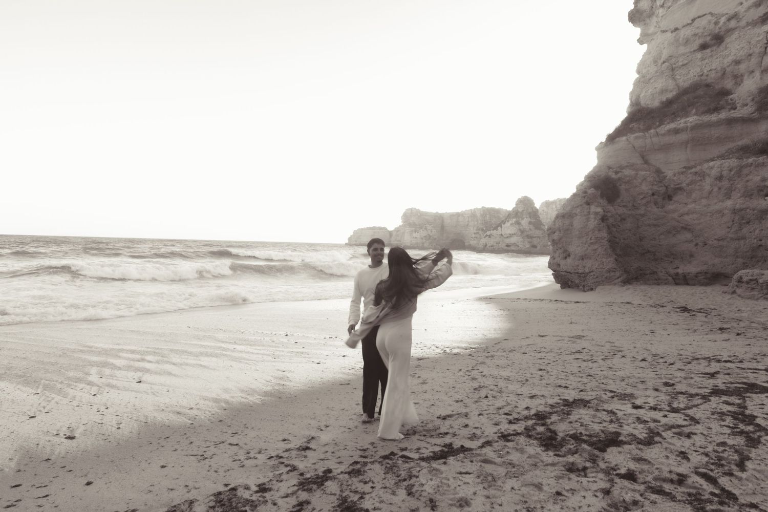 Couple embracing on a sandy beach near rocky cliffs in black and white photography.