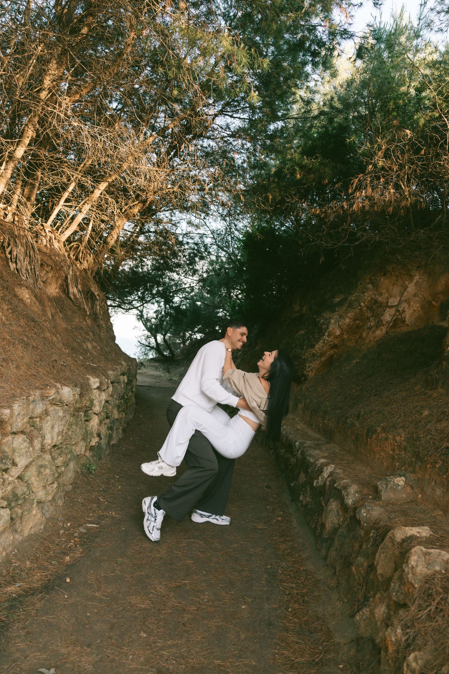 Romantic couple embracing on a scenic stone-walled pathway surrounded by lush trees outdoors.