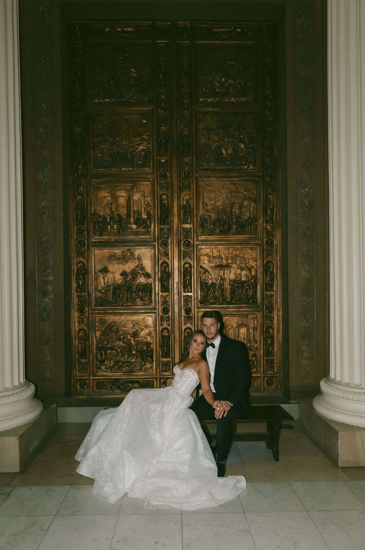 Elegant wedding couple poses in front of ornate carved wooden panels in a grand historic building interior.