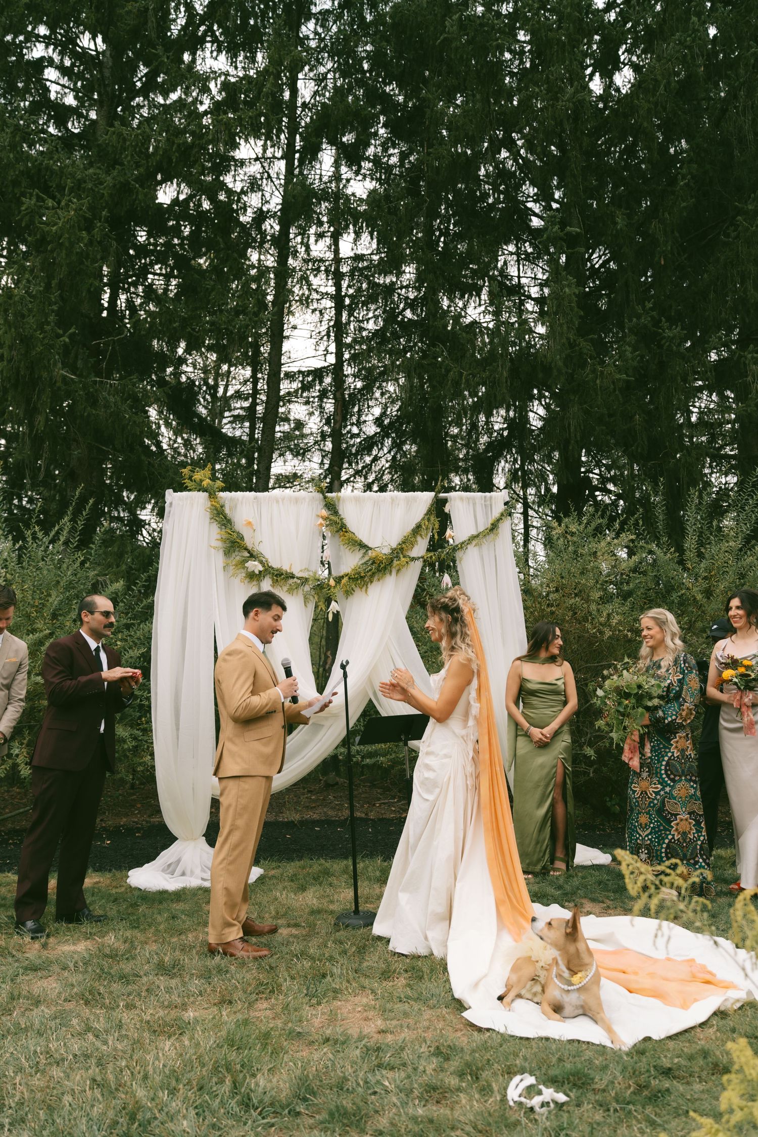Outdoor wedding ceremony setup with white drapes and greenery garland decorating the arch in a wooded setting.