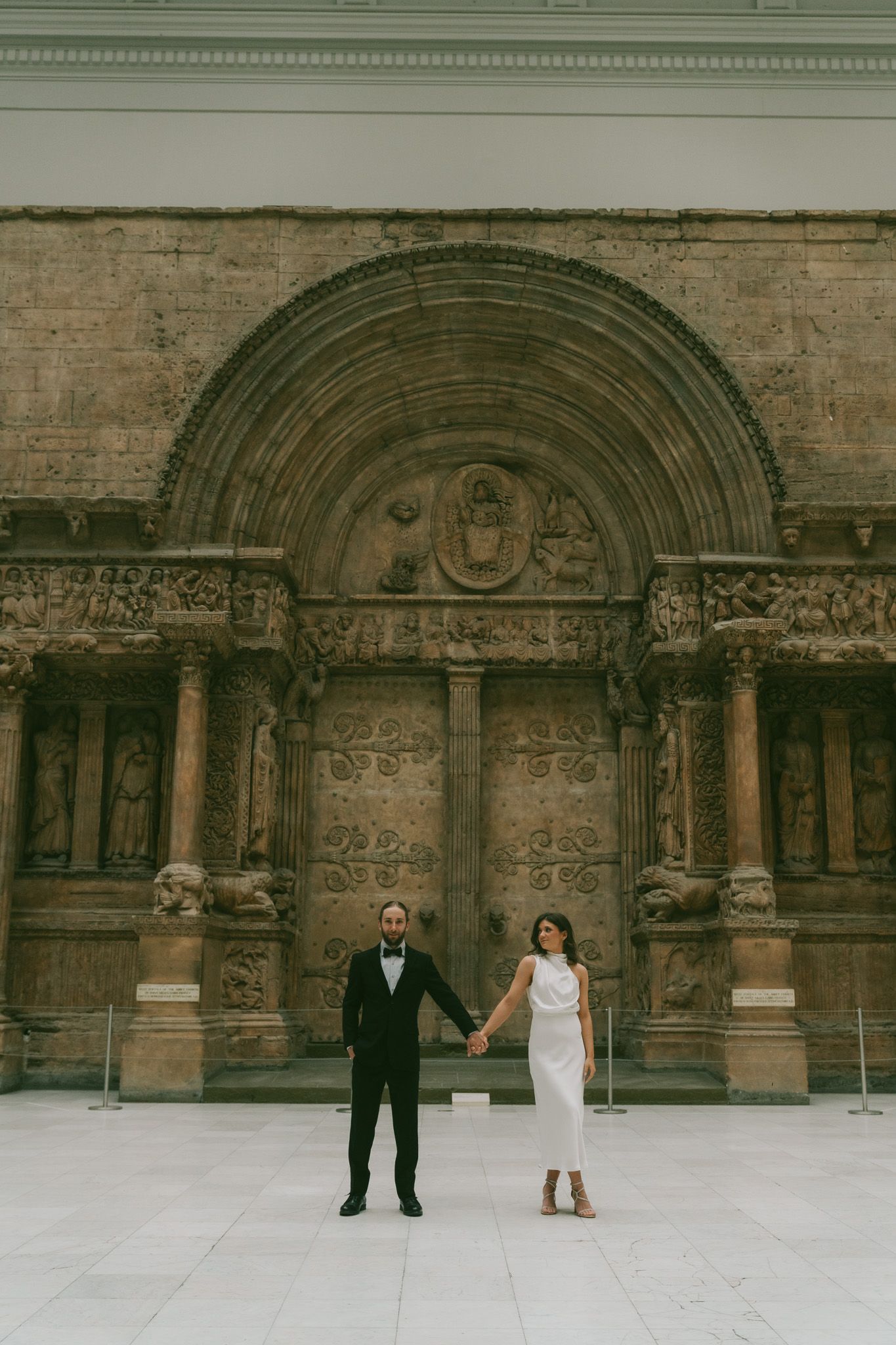 A couple holds hands while standing in front of an ornate stone church entrance with archways and detailed carvings in carnegie museum.