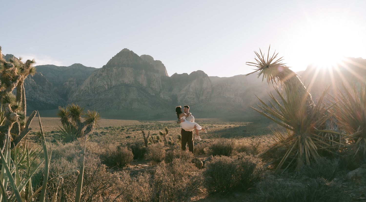 A couple embraces in a desert landscape at Red Rock Canyon during golden hour with mountains and yucca plants in view.