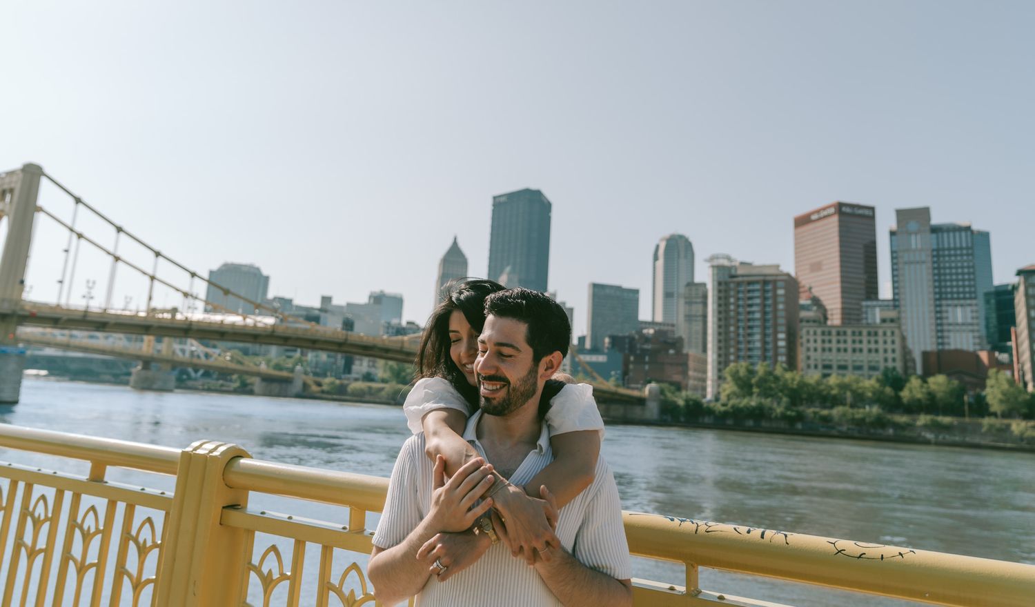 A couple embrace on a yellow bridge railing with Pittsburgh's downtown skyline and river in the background in north shore