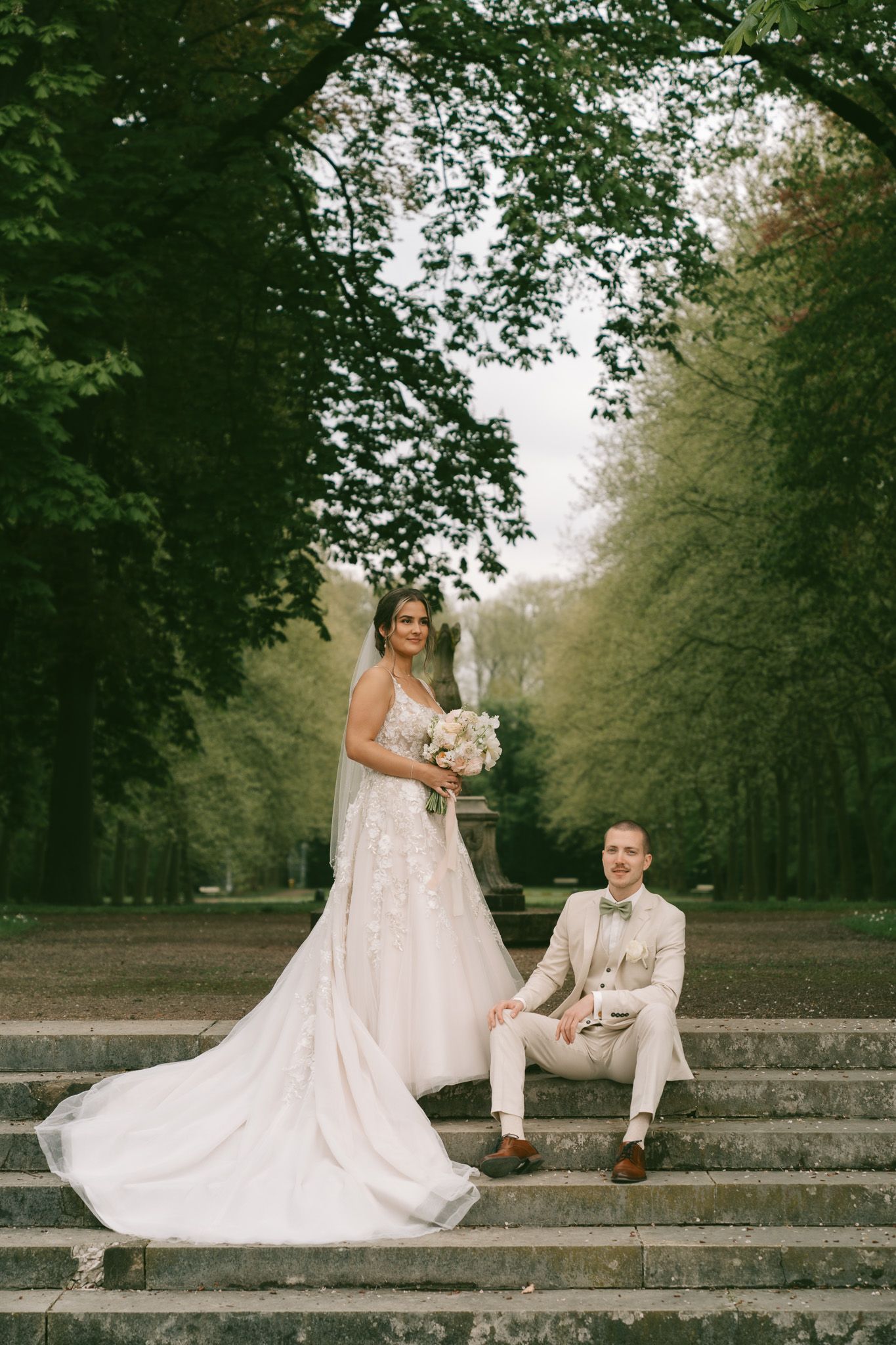 A wedding couple poses elegantly on stone steps surrounded by lush green trees in Germany.