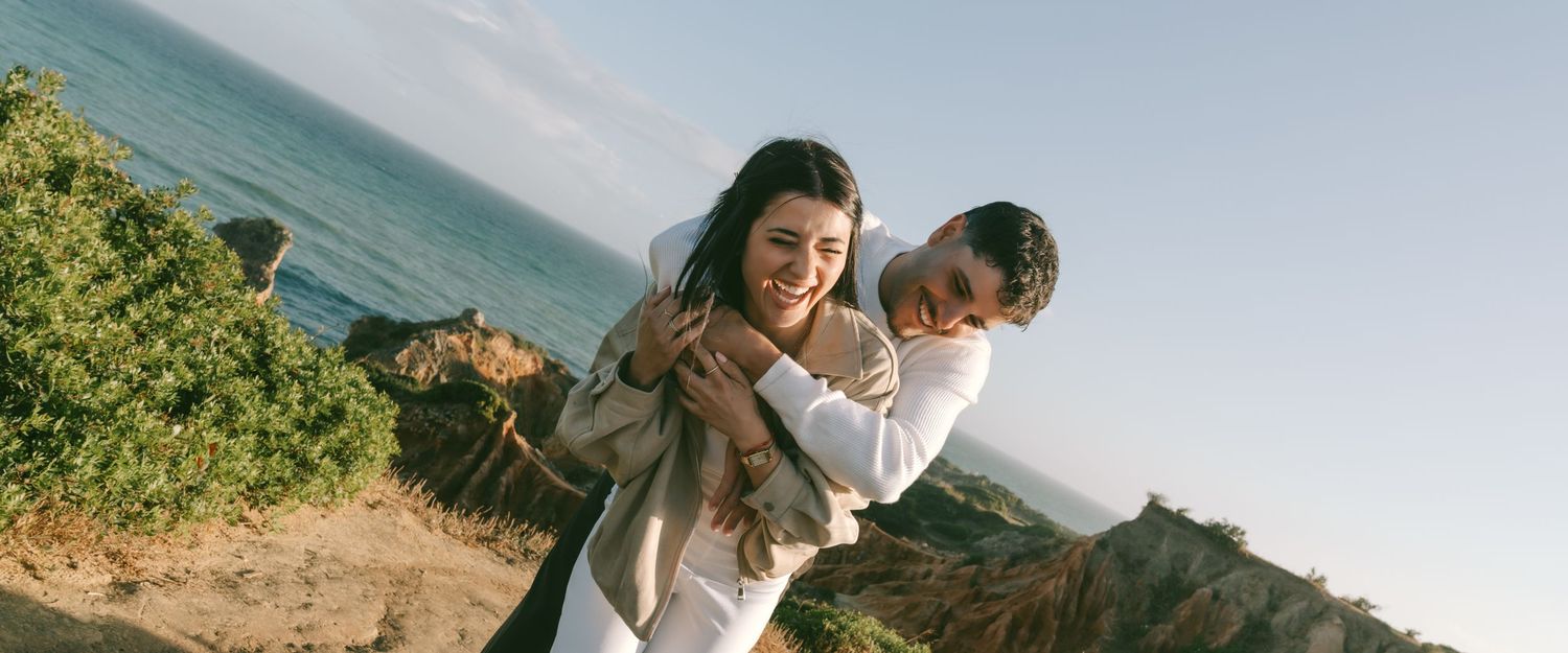 Couple sharing a joyful piggyback ride on a scenic coastal cliff with ocean views in golden hour light.