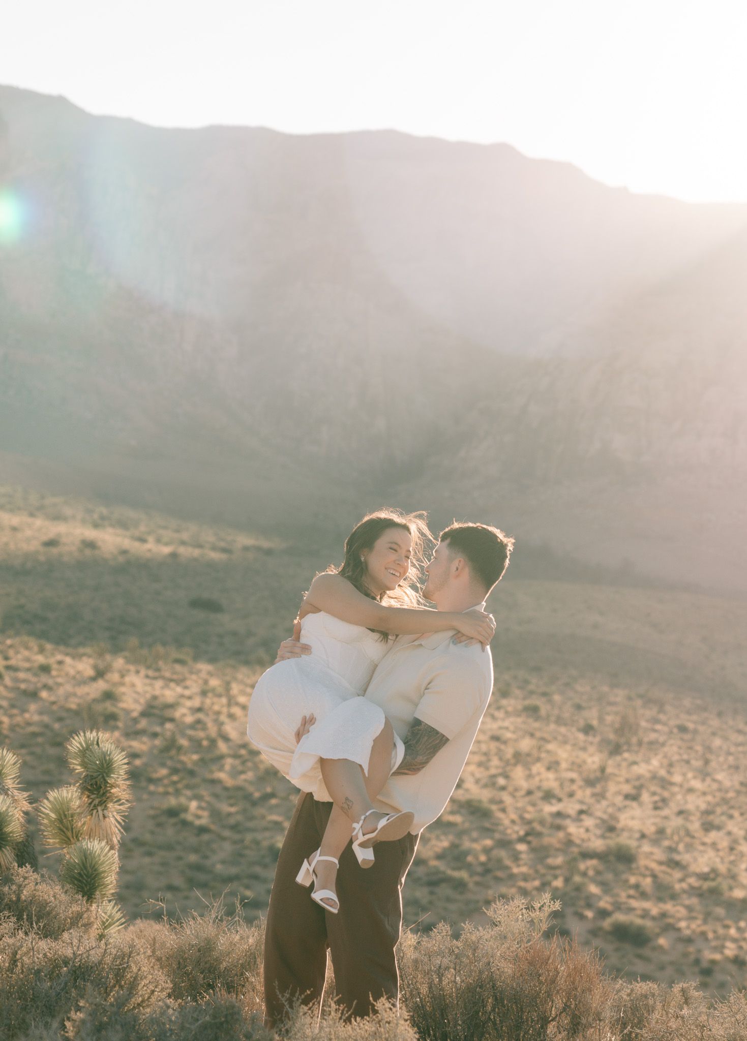 Couple embraces in the desert during golden hour with sun flare and Joshua trees in background in Joshua Tree, CA.