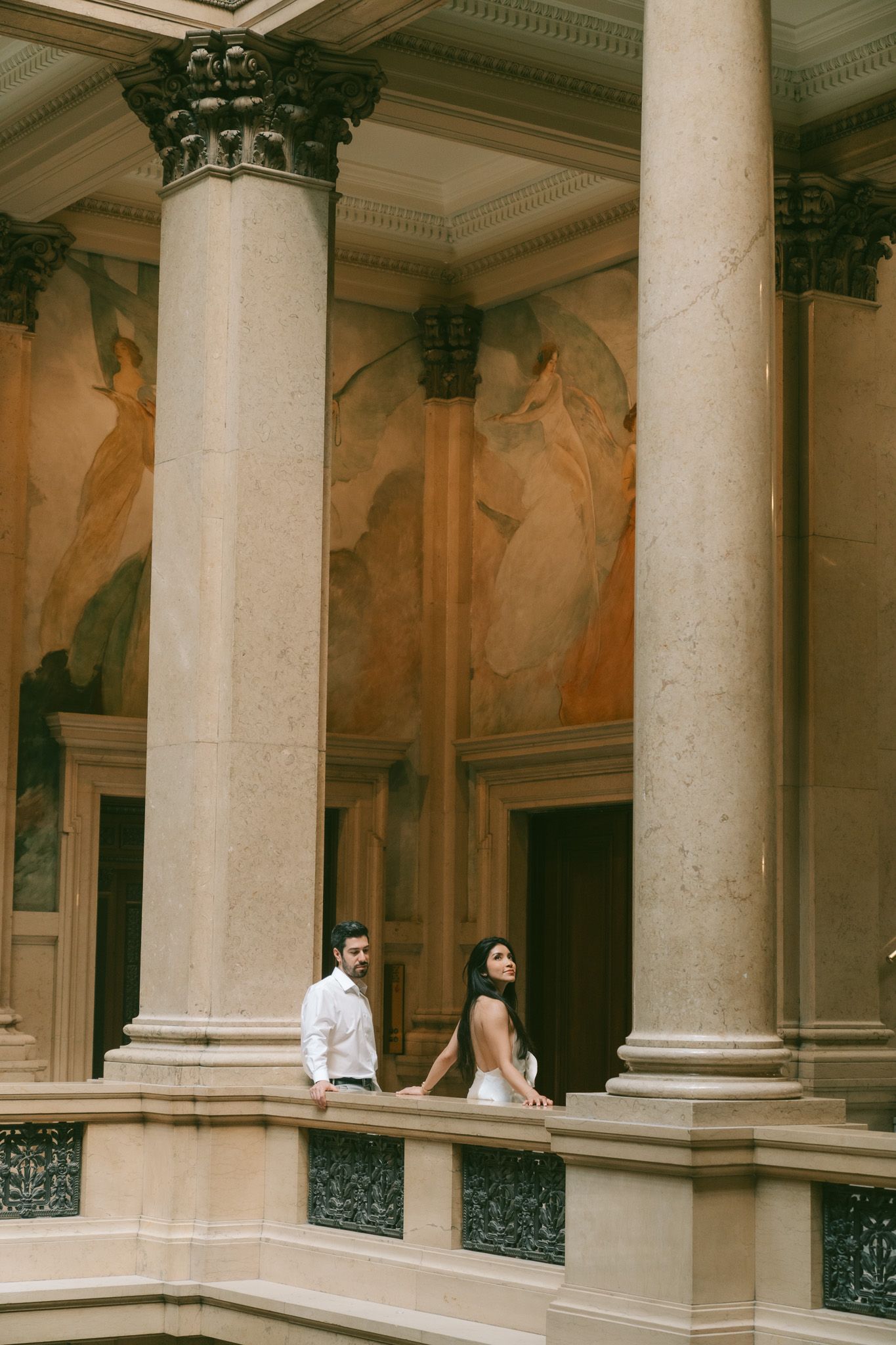 A couple poses elegantly between classical columns in a historic building.