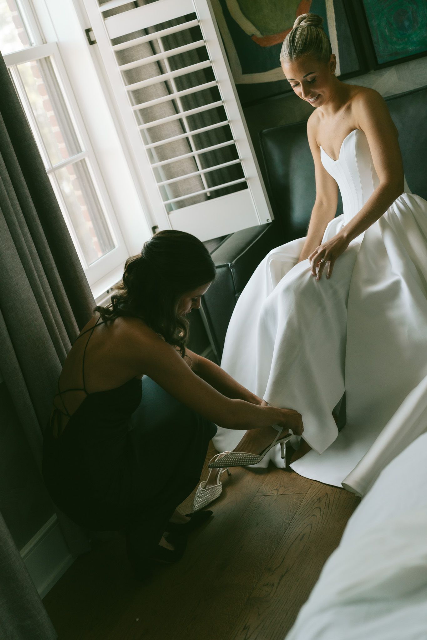 A person in dark clothing adjusts the hem of a flowing white wedding dress in a sunlit room.