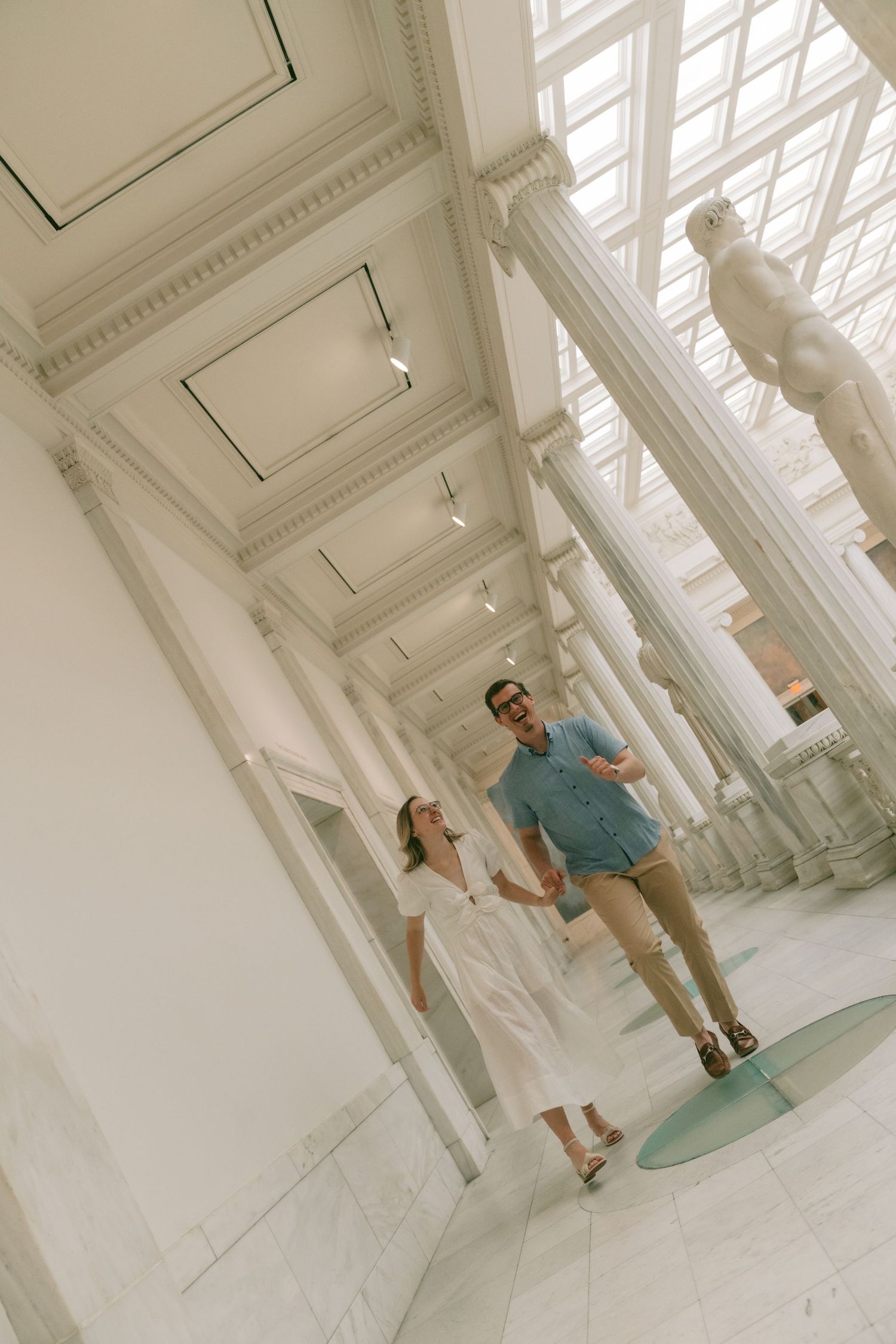 A couple walking through a bright white gallery space with tall columns and natural lighting.