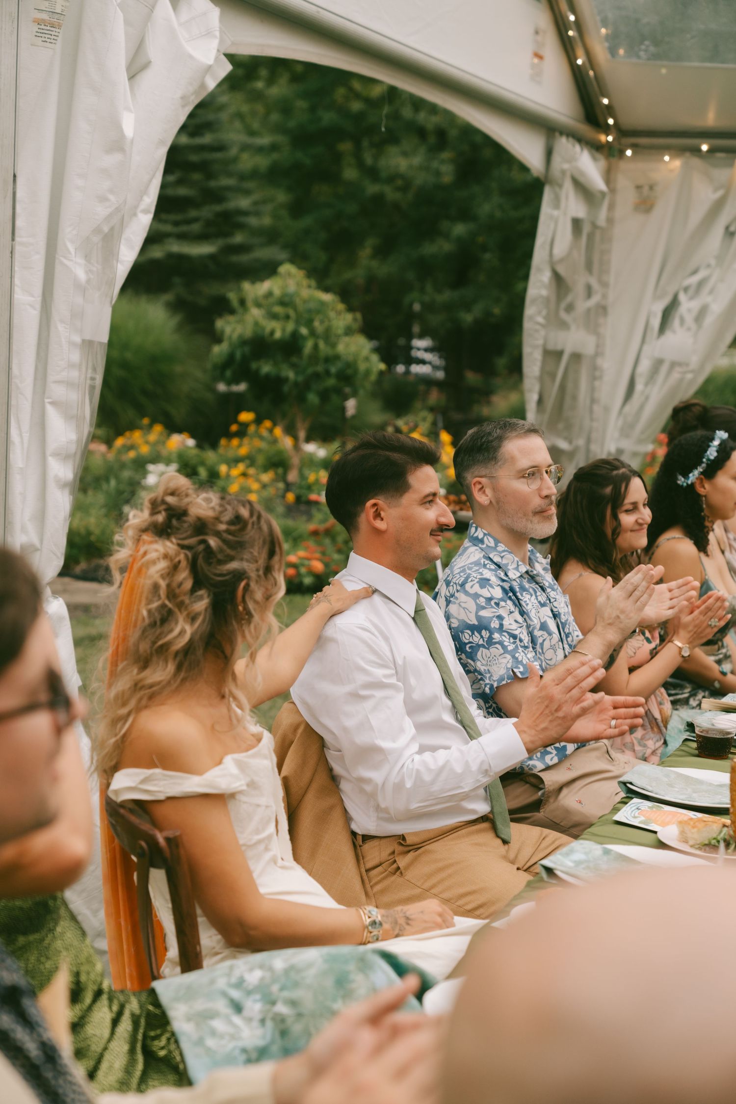 Wedding guests seated under white tent canopy applaud during outdoor celebration.