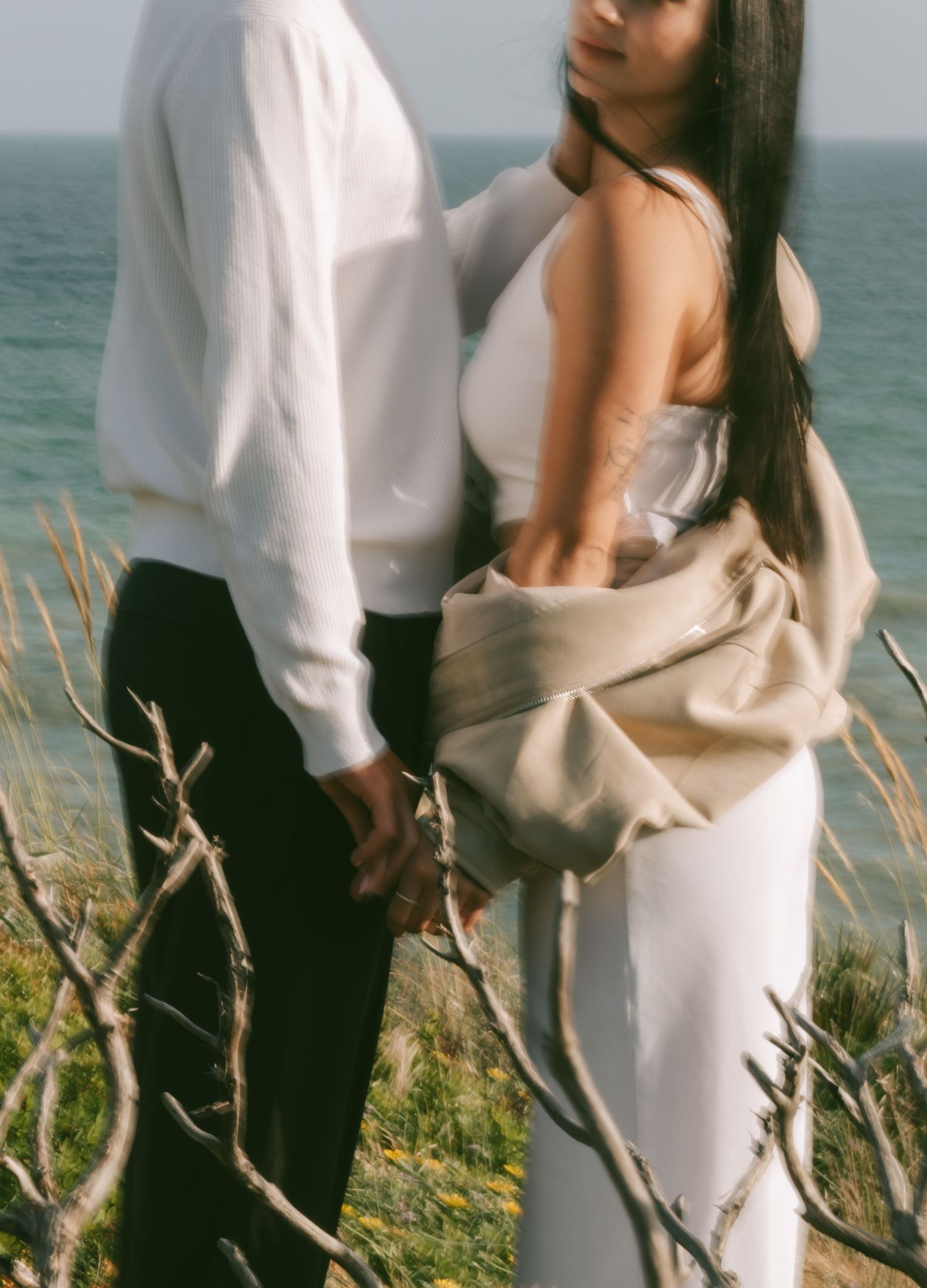 Couple holding hands by the ocean, woman in white dress with beige wrap, man in white shirt and black pants.