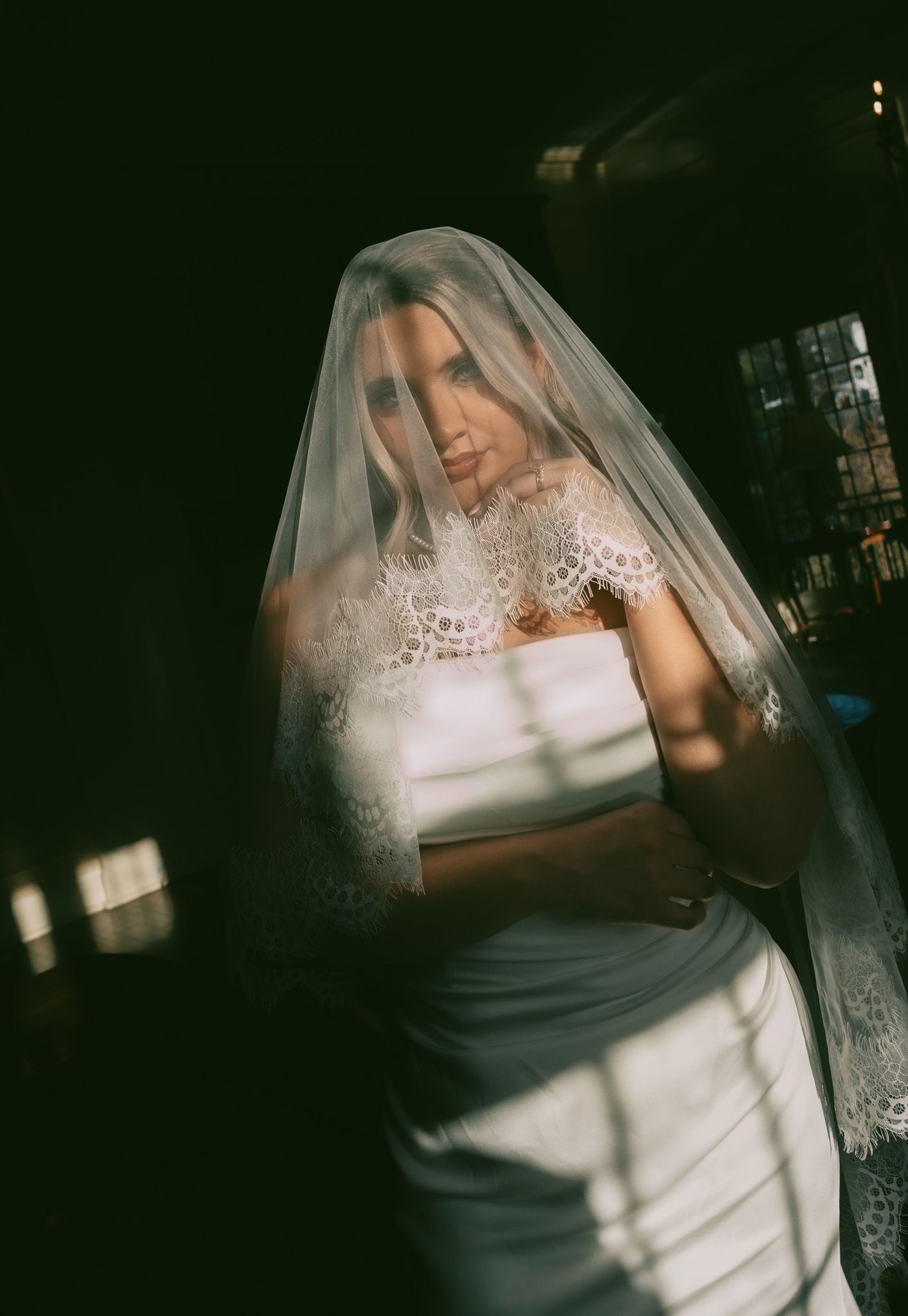 A bride in a classic white lace wedding dress and long veil poses for a moody, atmospheric portrait.