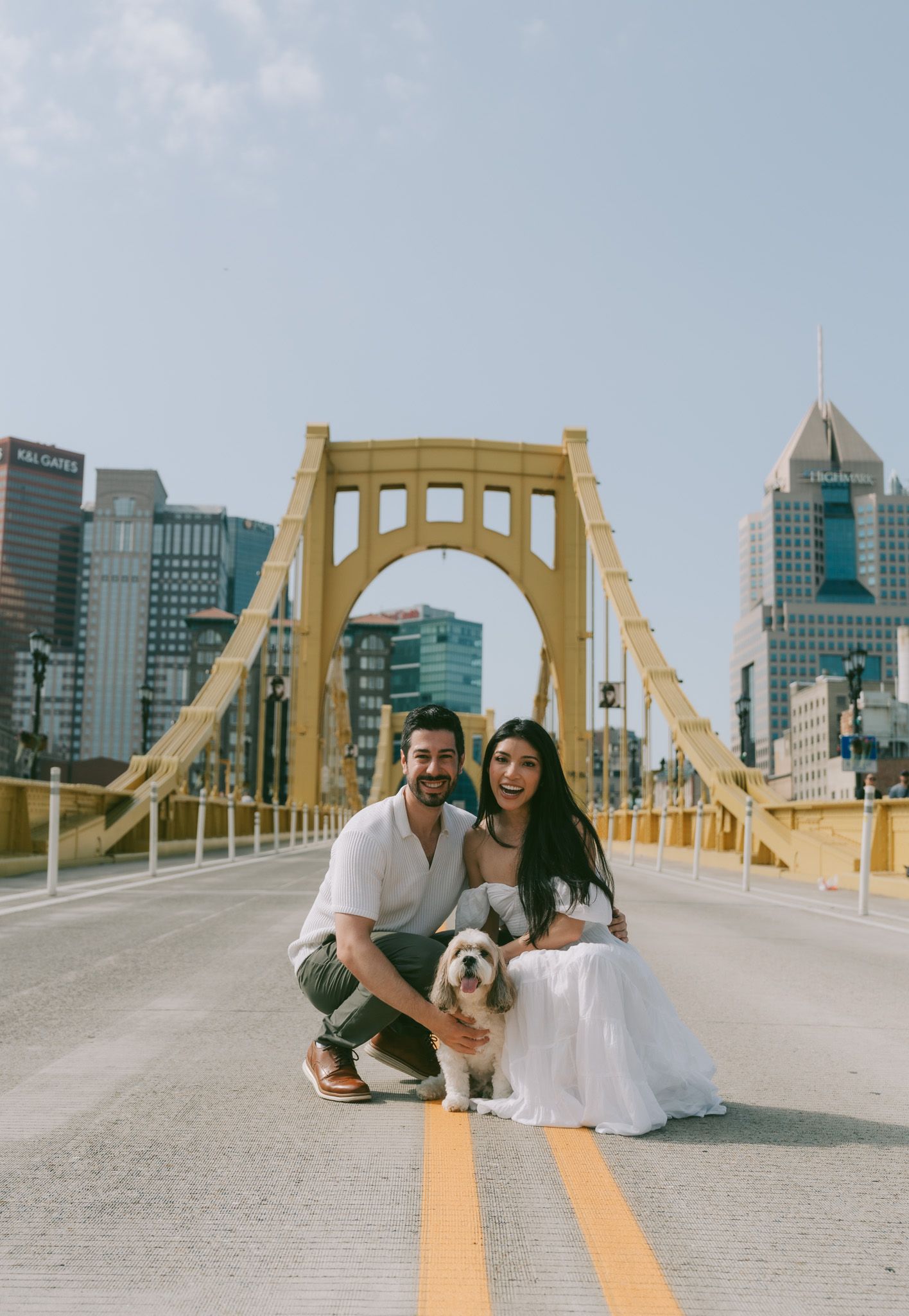 couple posing with their white dog on Pittsburgh's iconic yellow Roberto Clemente Bridge in north shore
