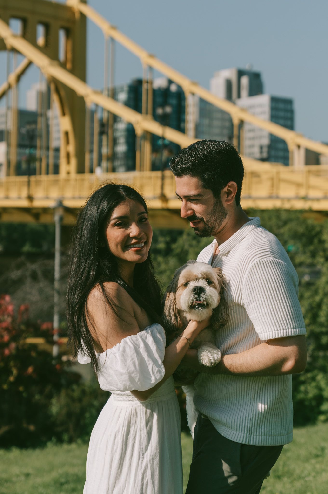 A couple stands with their small white dog near a yellow bridge in an urban park setting on a sunny day in north shore, pittsburgh