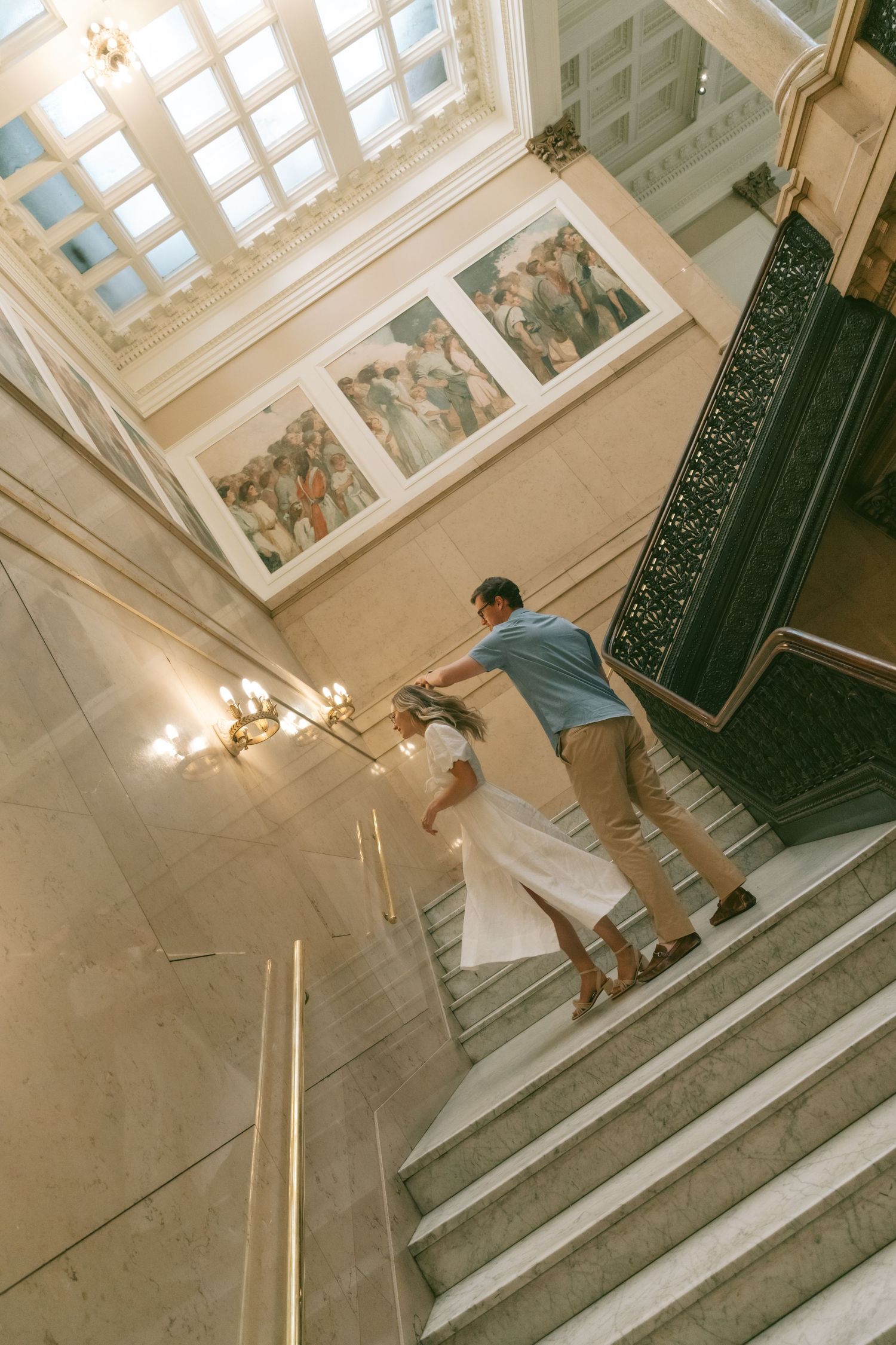 Sunlit marble staircase with ornate black railing and tall windows casting dramatic shadows in a historic building.