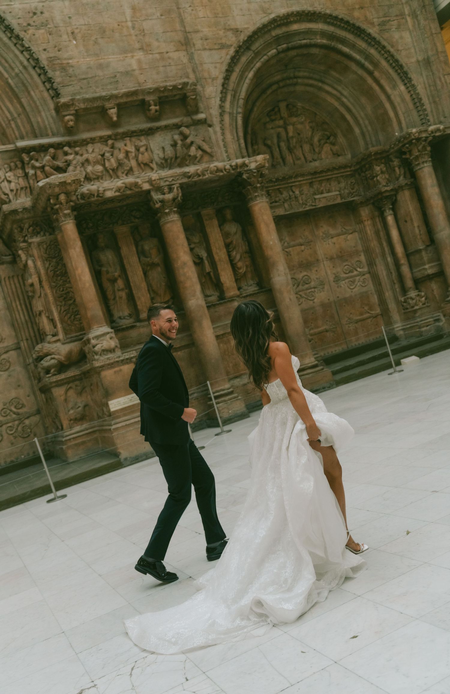 Wedding couple walks across a stone courtyard with historic architectural backdrop featuring arches and columns.