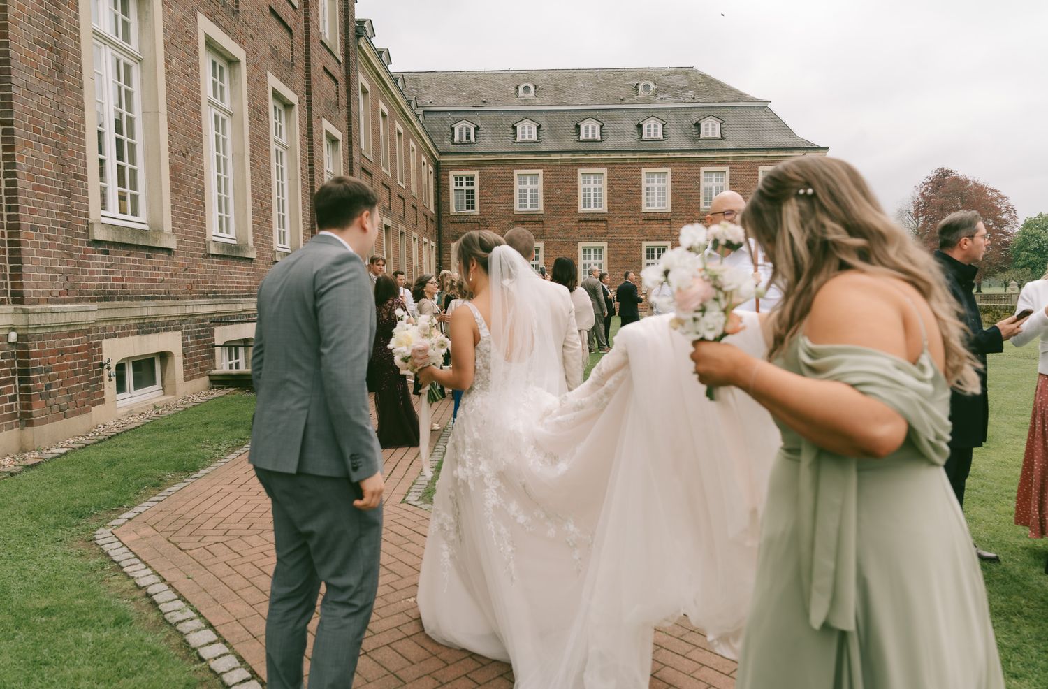 Wedding party helps adjust bride's flowing veil on brick pathway outside historic building in Germany.