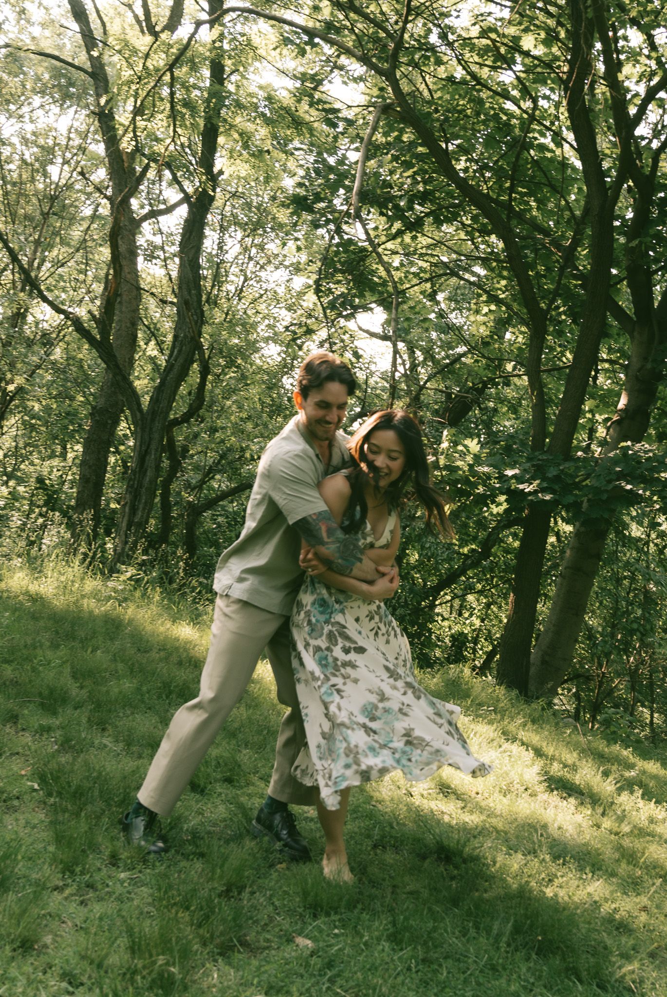 A couple shares a playful moment dancing together in a sunlit wooded area on a summer day at shenley park, pittsburgh.