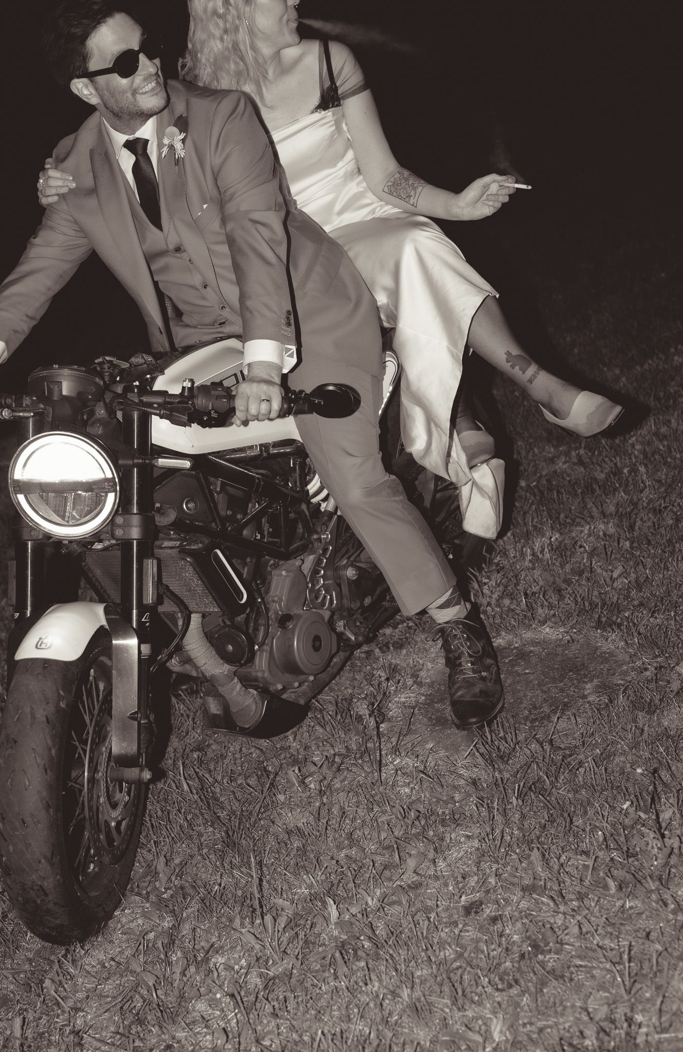 Stylish couple posing on vintage motorcycle in wedding attire at night.