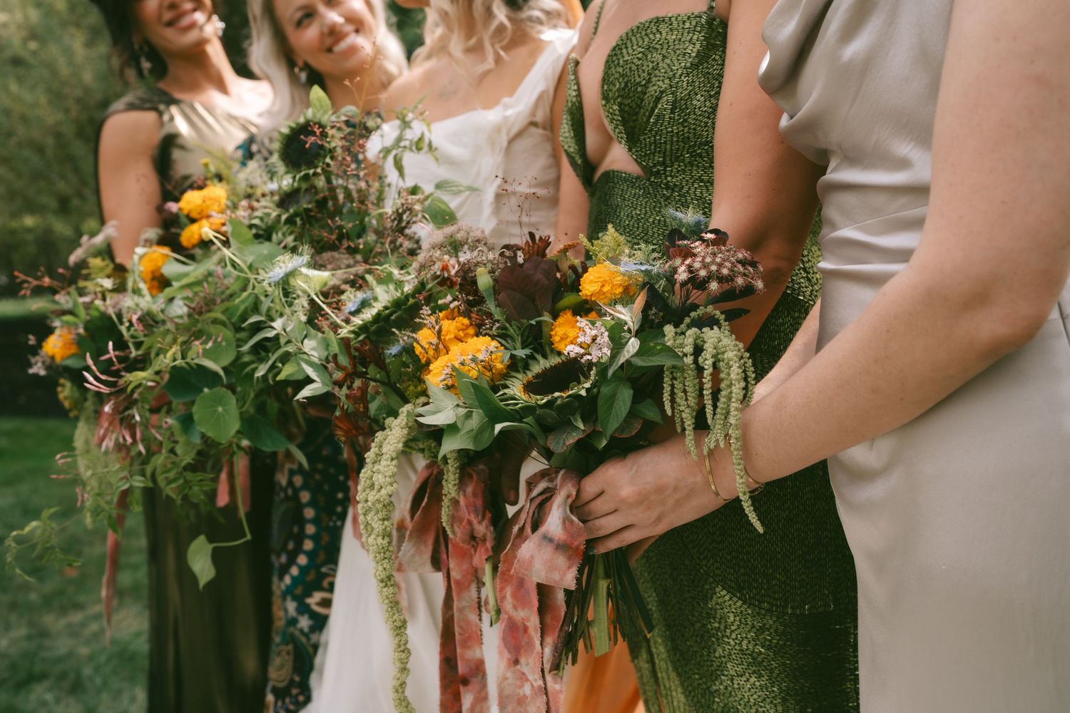 Wedding party members hold vibrant bouquets with orange flowers and greenery against emerald green dresses at outdoor ceremony.