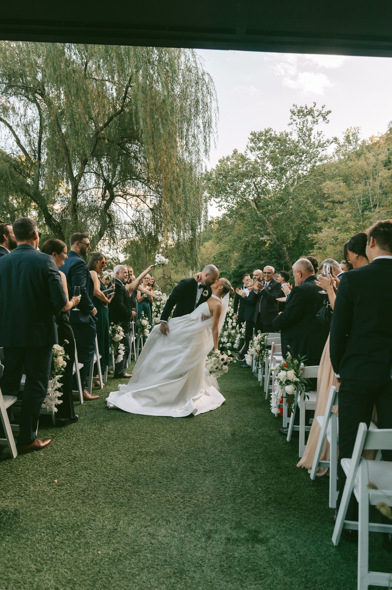 Wedding guests stand and take photos as newlyweds walk down the aisle at an outdoor garden ceremony under willow trees.