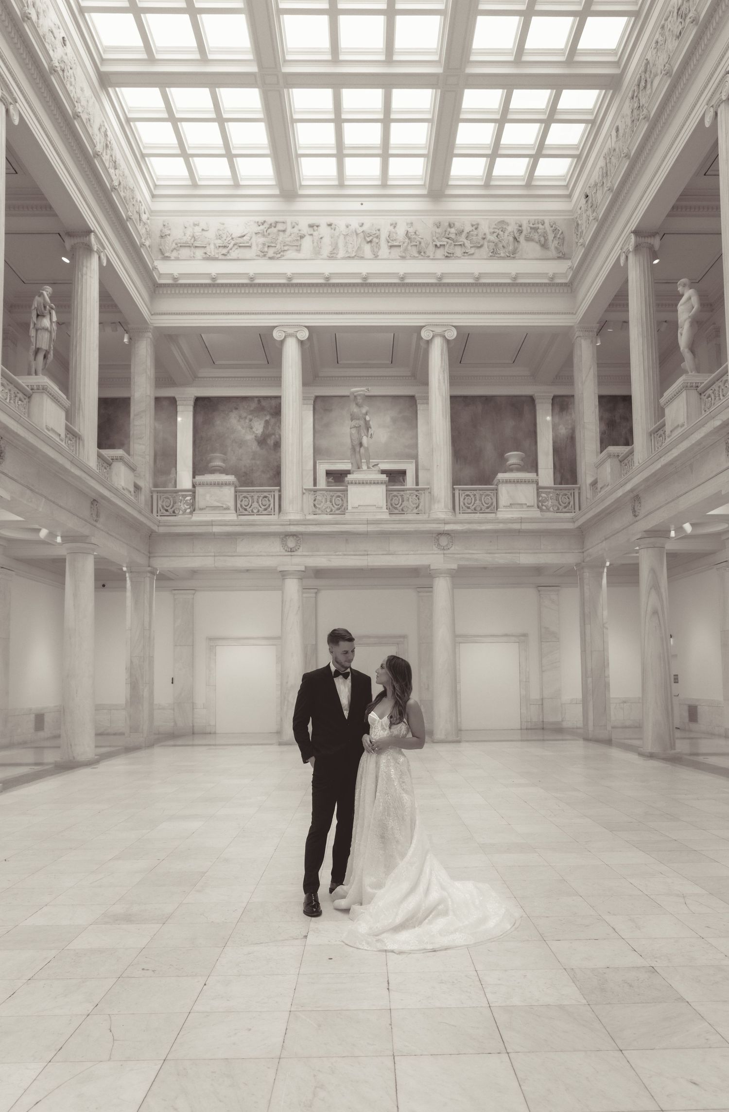 A romantic wedding couple stands in an elegant white marble ballroom with skylights and classical architectural details.