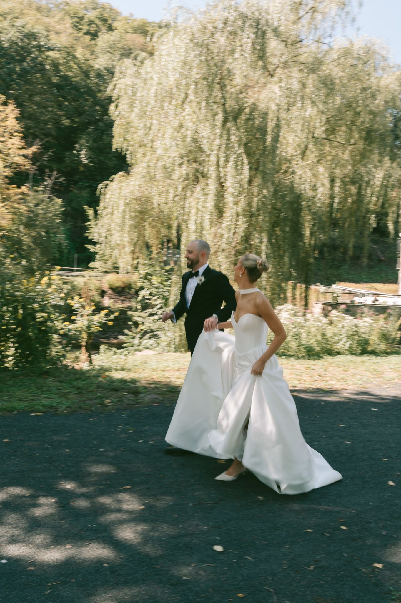 A bride in a flowing white dress walks through a garden setting with graceful weeping willow trees in the background.